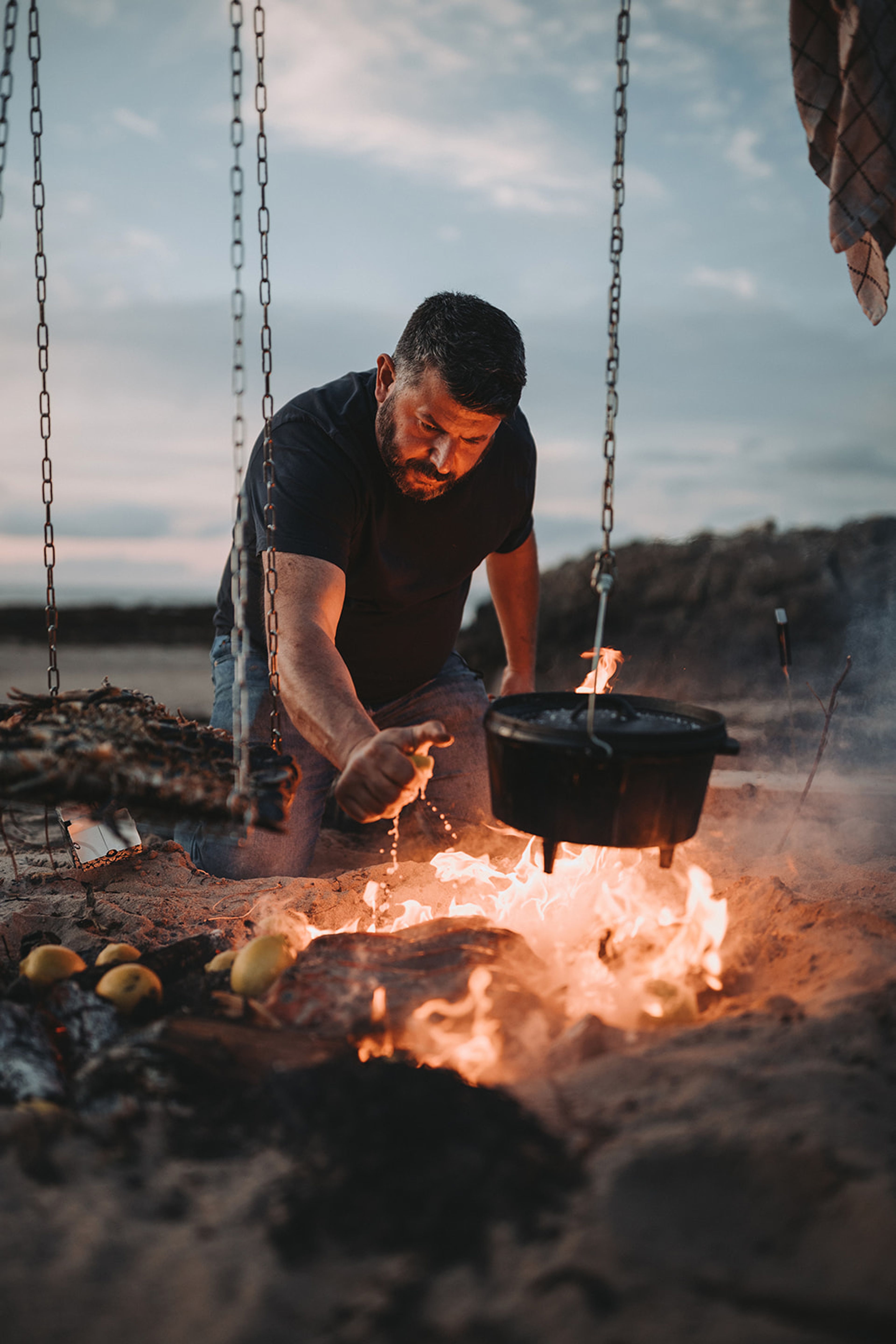 Chef squeezing lemon over a beach campfire at dusk, cast-iron pot hanging above the flames