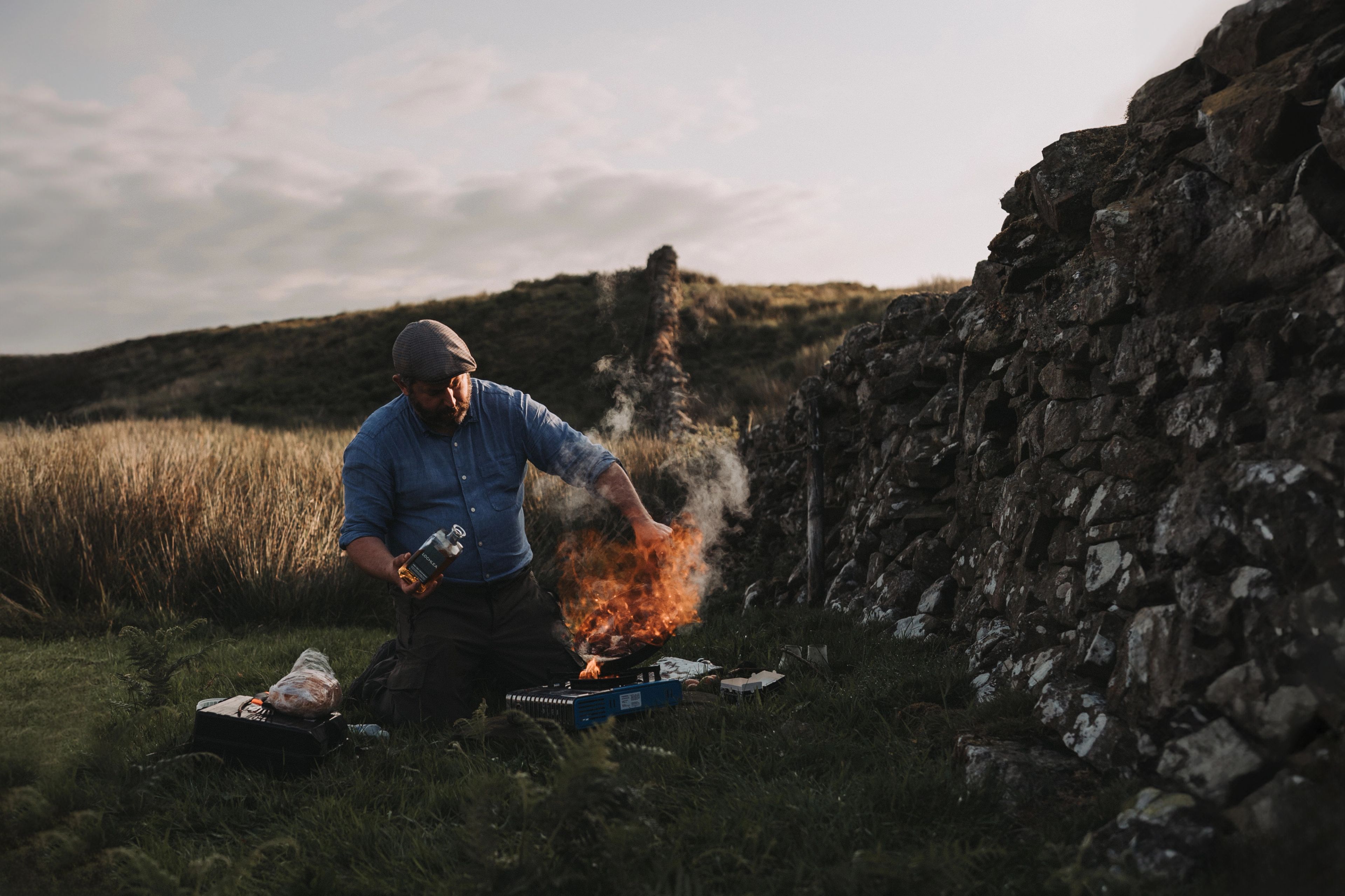 Man cooking over an open flame beside a drystone wall at dusk, outdoor lifestyle photography