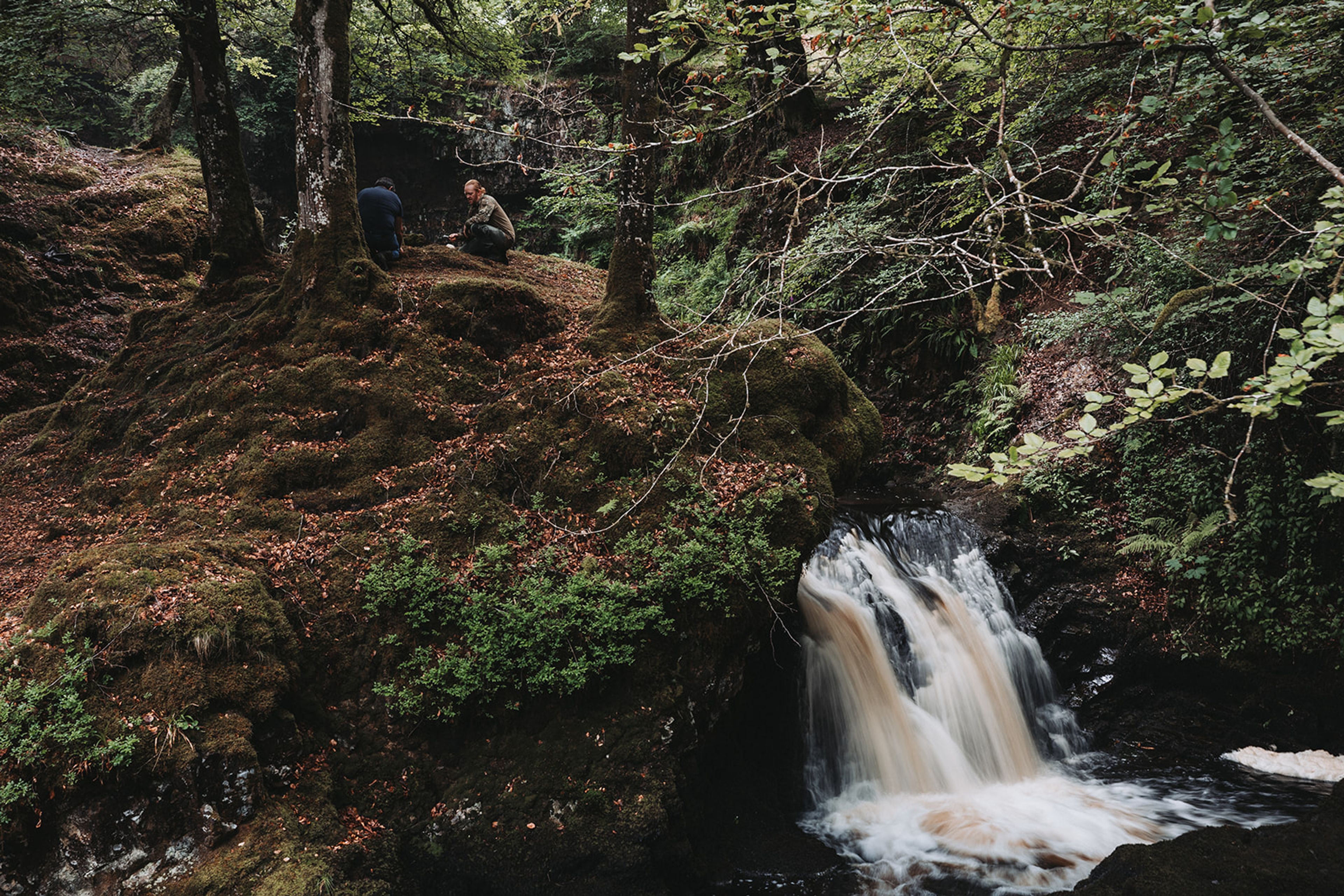 Waterfall in a mossy wooded glen near The Kirkmichael Arms, with two people crouched on the bank