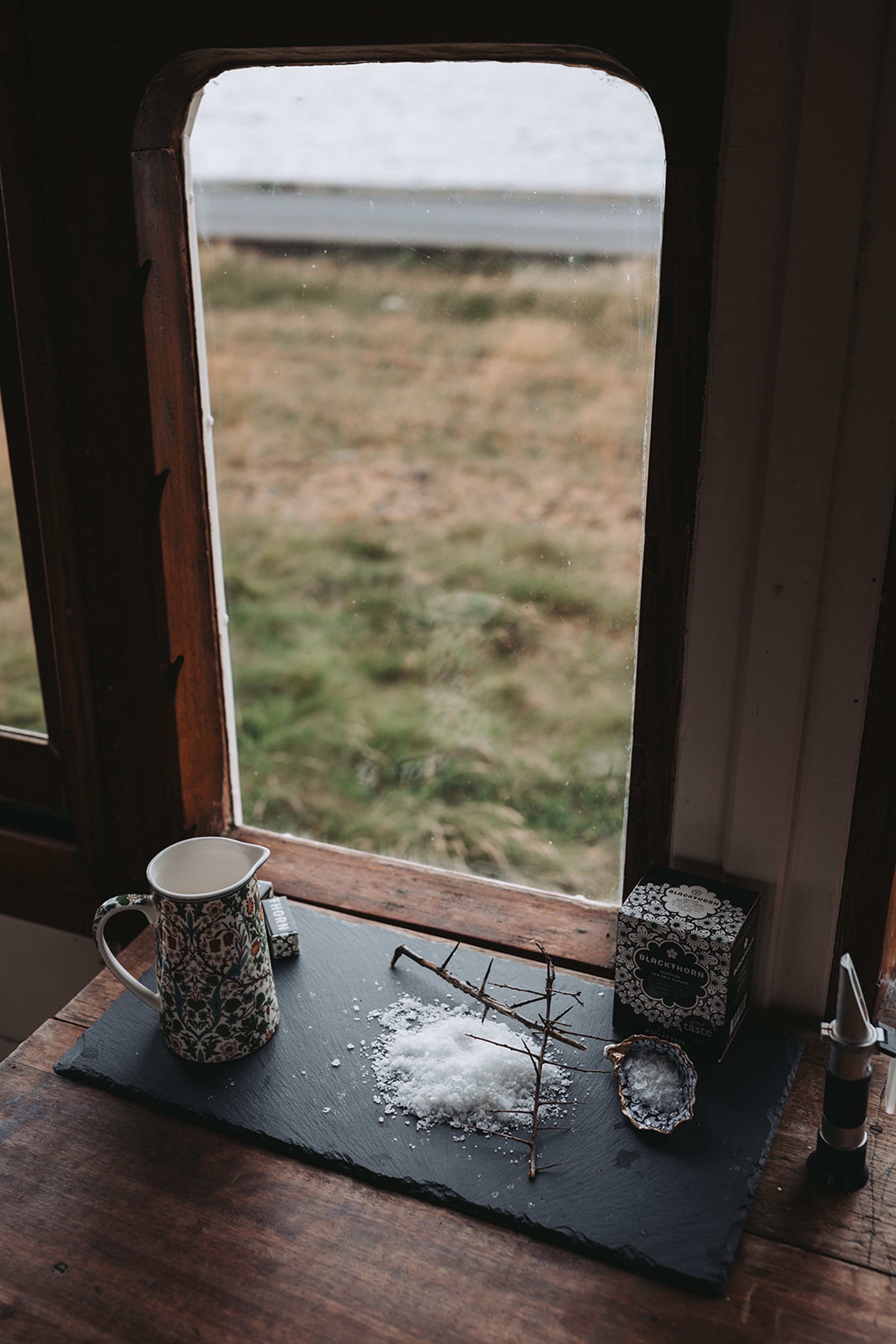 Sea salt, patterned jug and shell arranged on slate by a window overlooking coastal grassland