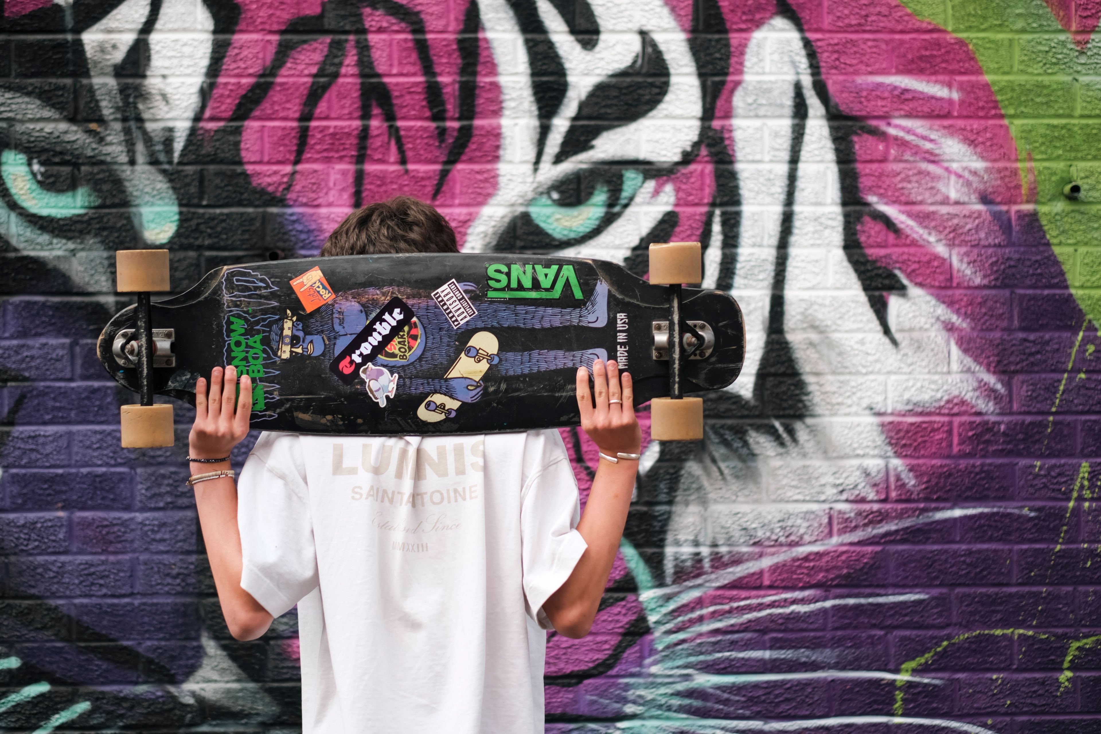 Teenager holding a sticker-covered skateboard behind his head in front of a tiger mural, from the BeU Swindon shoot