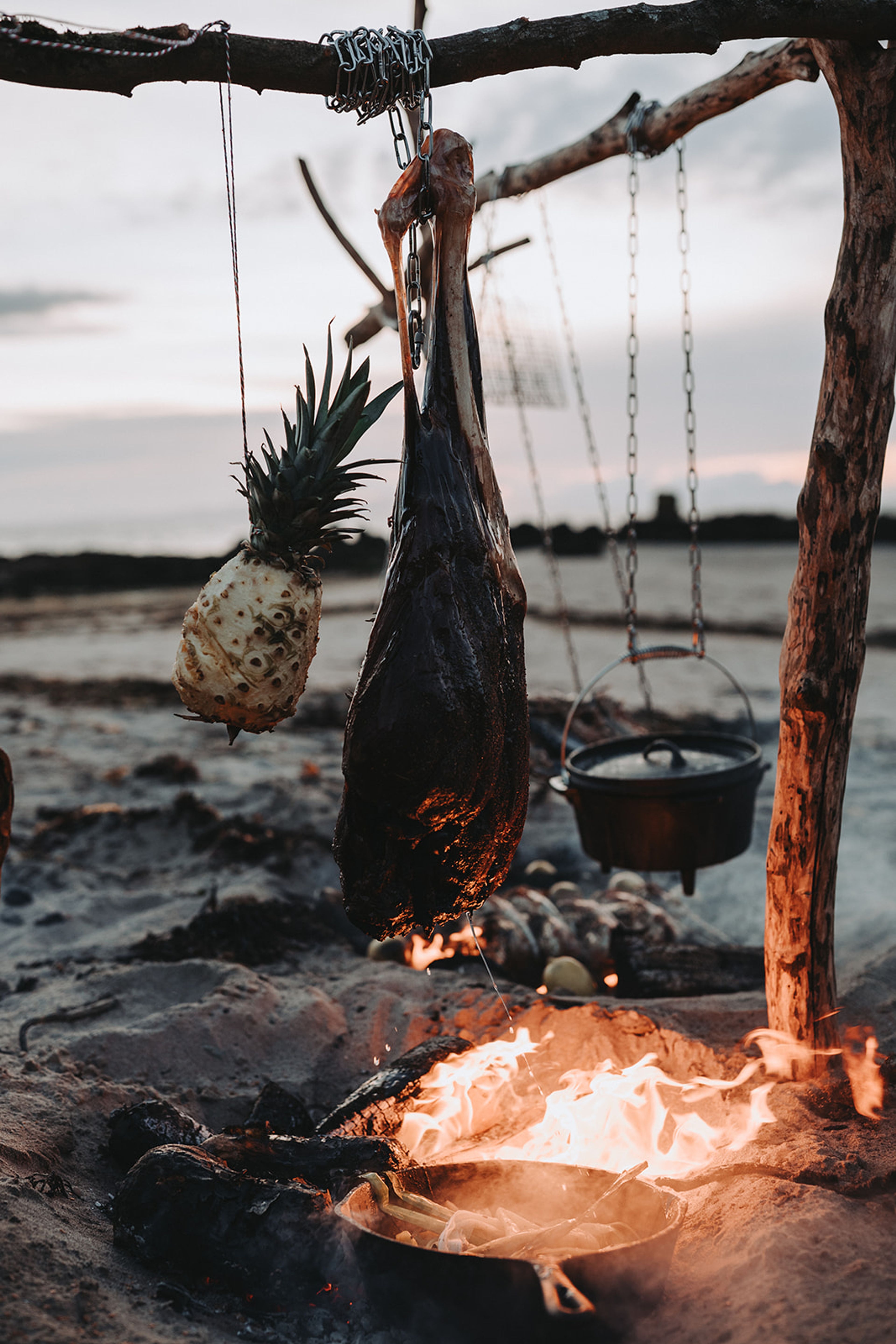 Cured meat and pineapple hanging from a driftwood frame over a beach fire, The Kirkmichael Arms food photography