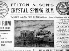 Newspaper advertisement for Felton & Son's Crystal Spring Rum, a copper-distilled molasses rum from South Boston, with an engraving of the company's waterfront distillery and "New England Rum" painted across its wall.