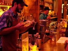 A bartender in an apron measuring a pour into a jigger behind a fairy-lit wooden bar, a second bartender working behind.