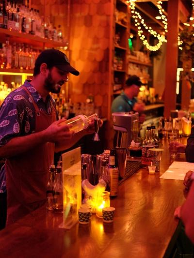 A bartender in an apron measuring a pour into a jigger behind a fairy-lit wooden bar, a second bartender working behind.