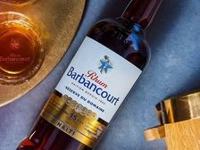 A bottle of Rhum Barbancourt Réserve du Domaine 15 ans laid on slate beside two branded glasses of amber rhum and small oak barrels.
