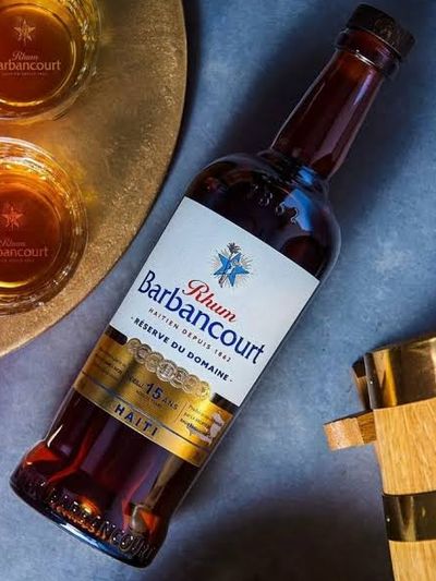 A bottle of Rhum Barbancourt Réserve du Domaine 15 ans laid on slate beside two branded glasses of amber rhum and small oak barrels.