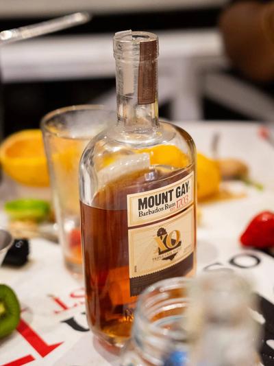 A bottle of Mount Gay XO on a bar mat crowded with a jigger, mixing glass and cut fruit part-way through building cocktails.