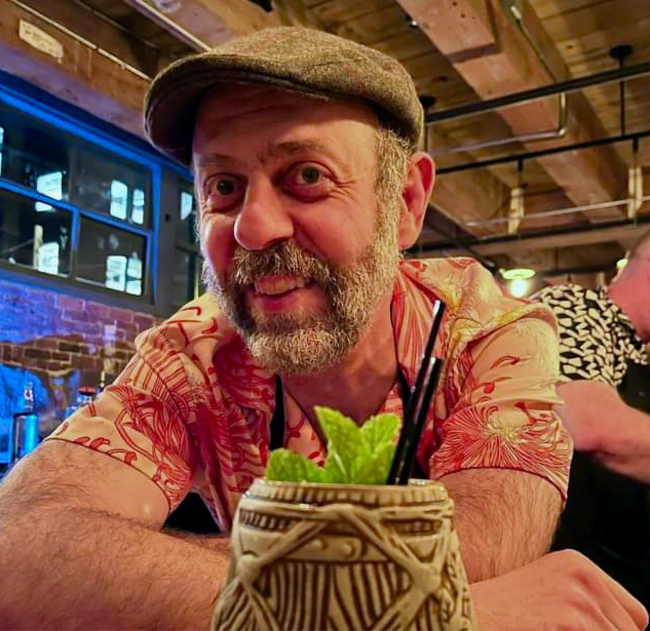 Frederic Yarm in a flat cap and patterned aloha shirt, leaning on a bar behind a carved tiki mug garnished with fresh mint.