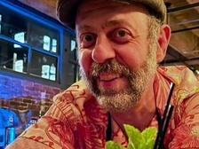Frederic Yarm in a flat cap and patterned aloha shirt, leaning on a bar behind a carved tiki mug garnished with fresh mint.