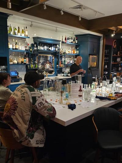 A presenter leads a seated tasting behind the white bar counter at Silver Girl, guests ranged around rows of tasting glasses and notes.