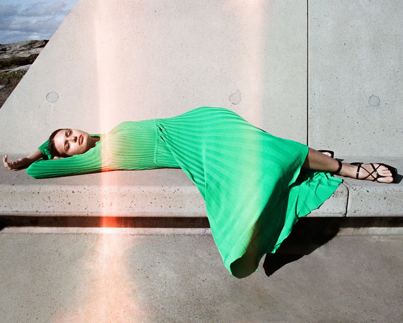 Brunette girl laying on concrete bench with her hands behind her head in green pleated dress. 