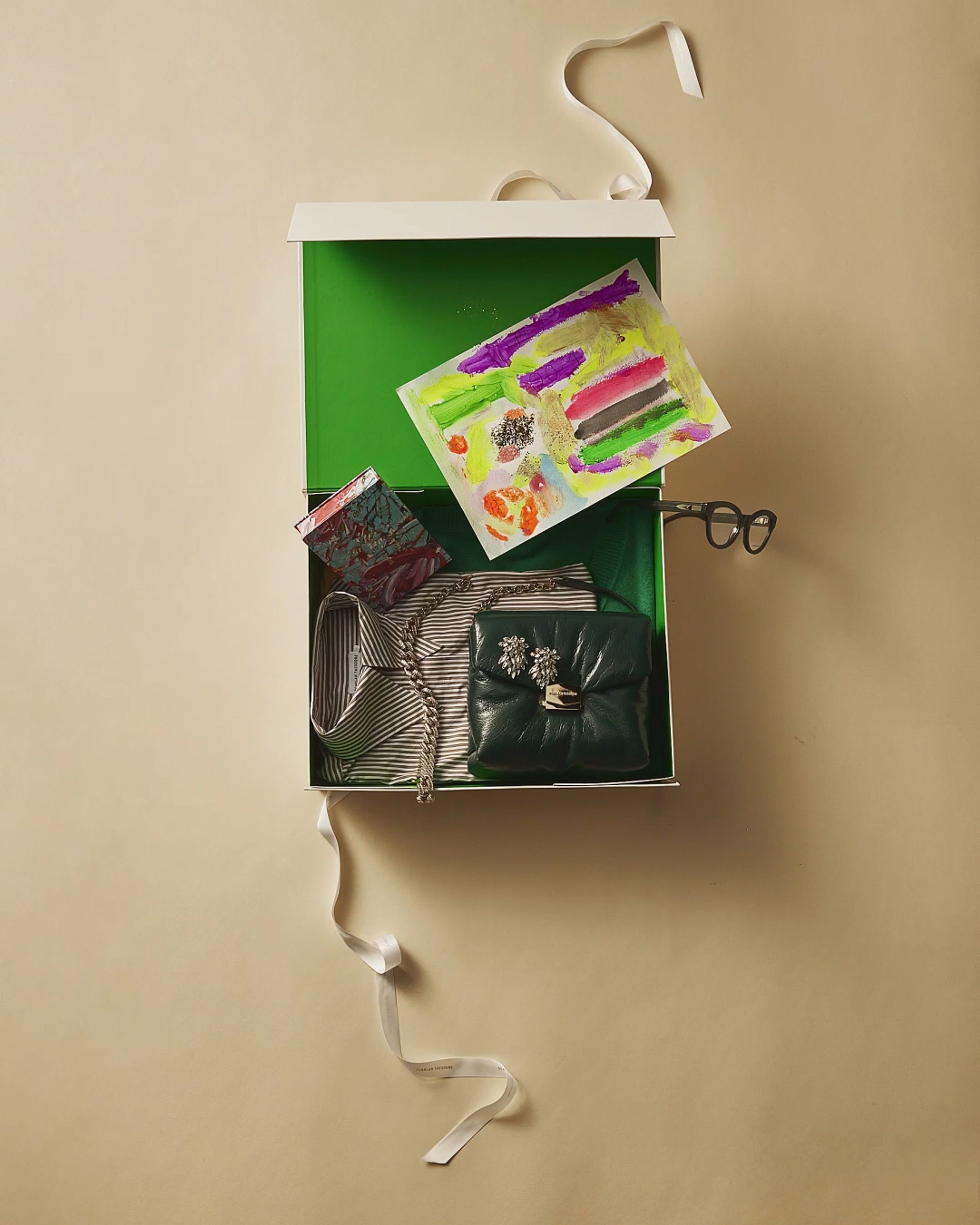 Flatlay of an open green gift box with green pillow bag, black and white striped shirt, and child's drawing