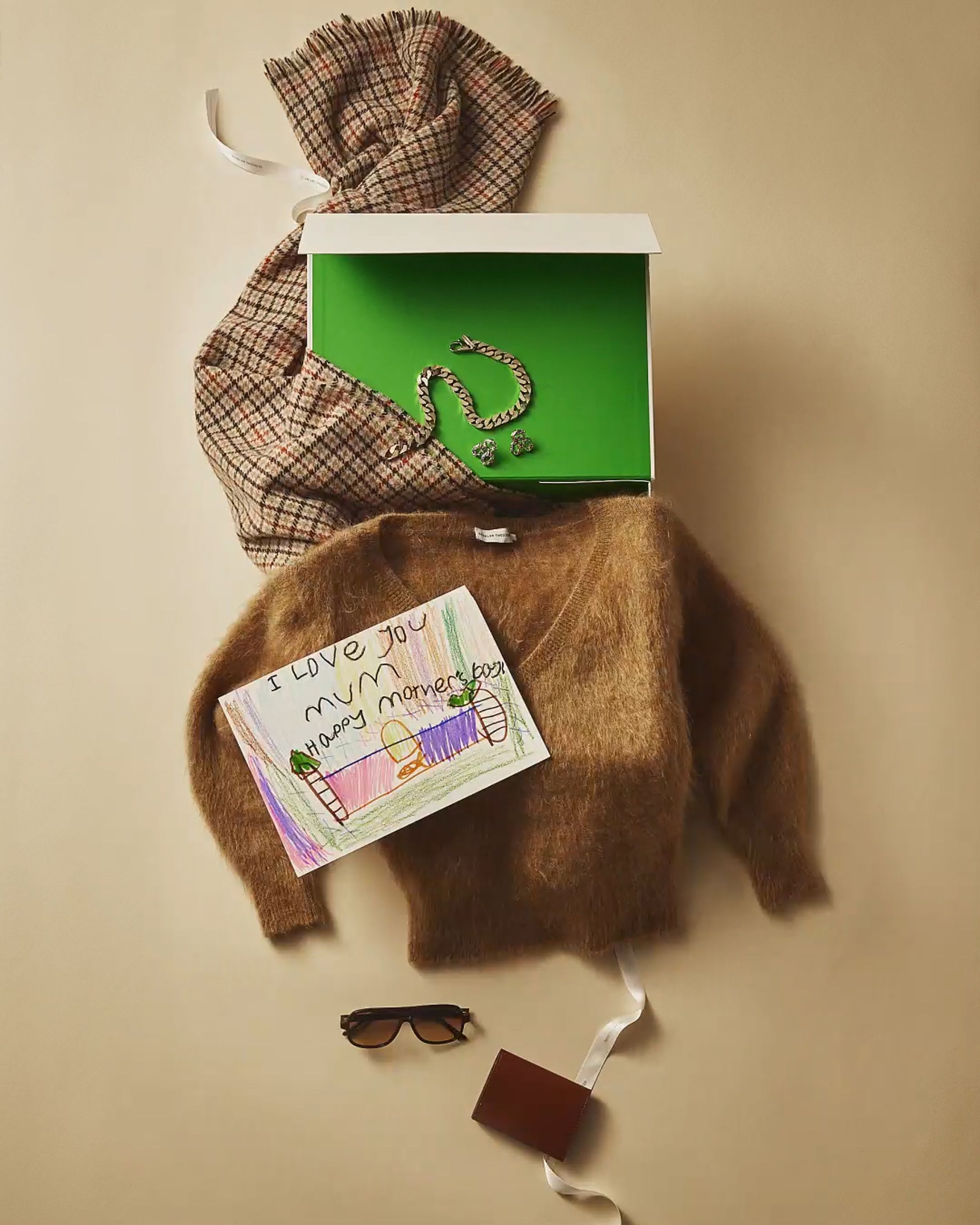 Flatlay of an open green gift box with khaki sweater, scarf, gold necklace and child's drawing