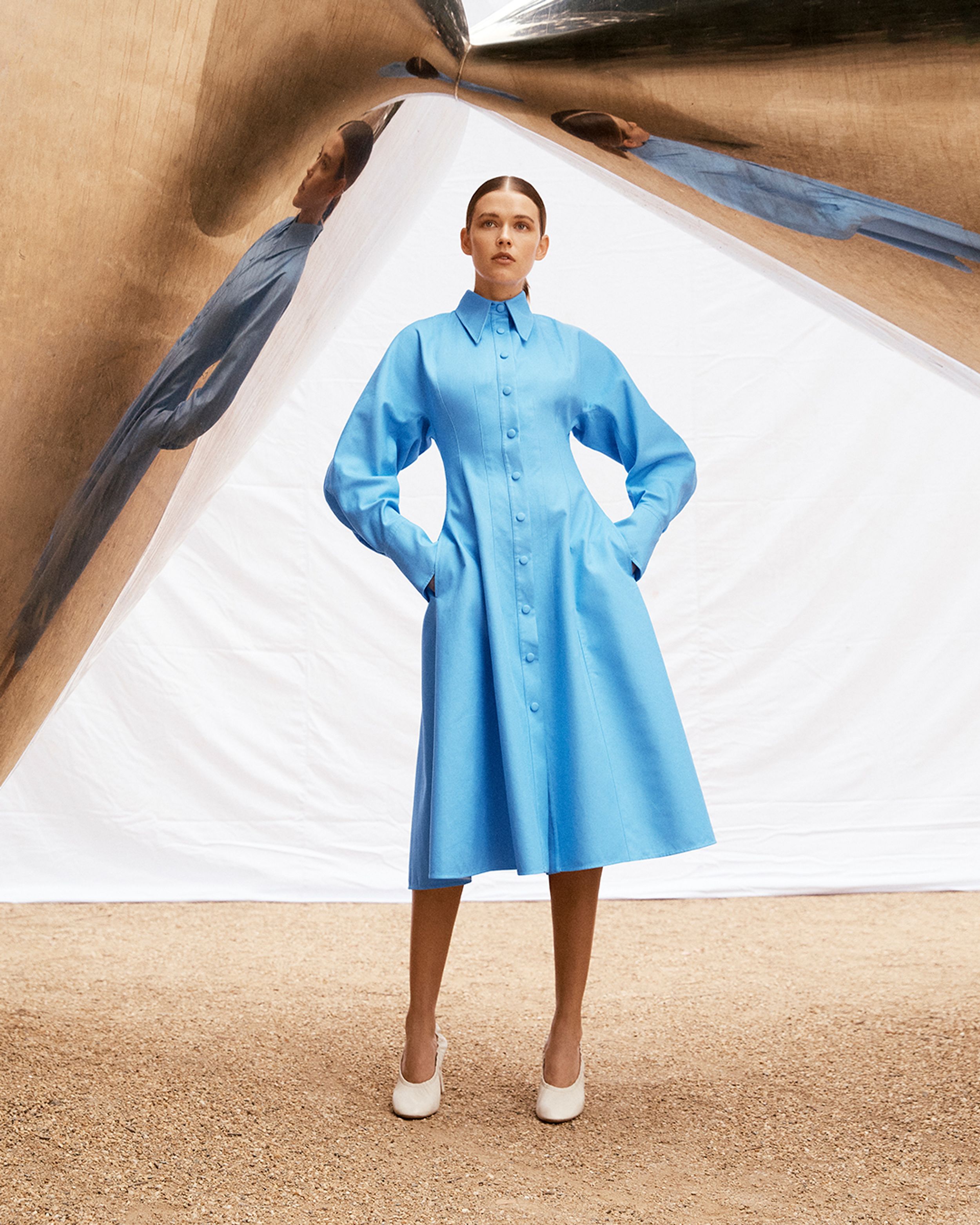 Brunette model standing under a mirrored sculpture wearing a blue shirt dress