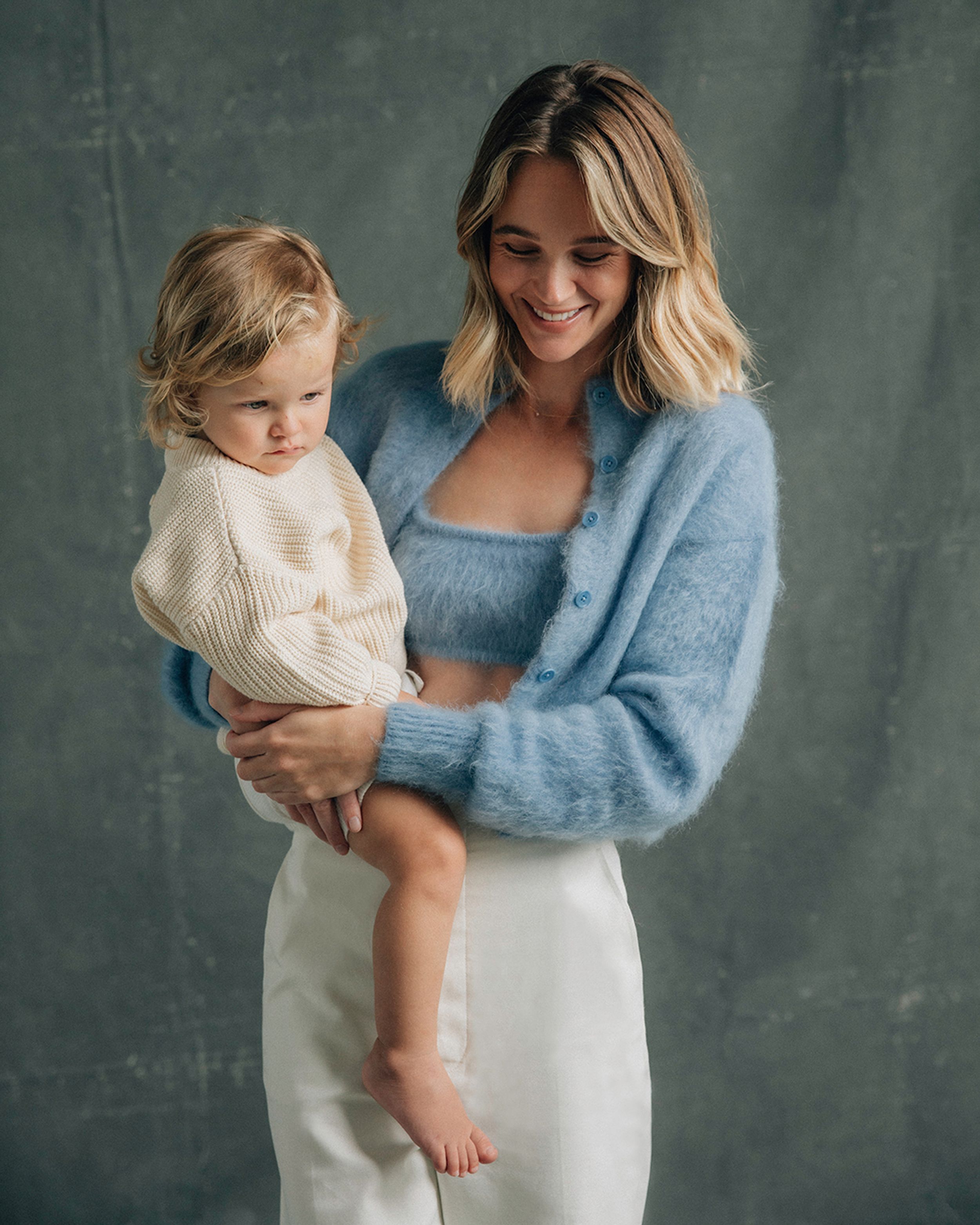 Rosie Tupper looking down smiling wearing a blue mohair cardigan and white trousers while holding her son