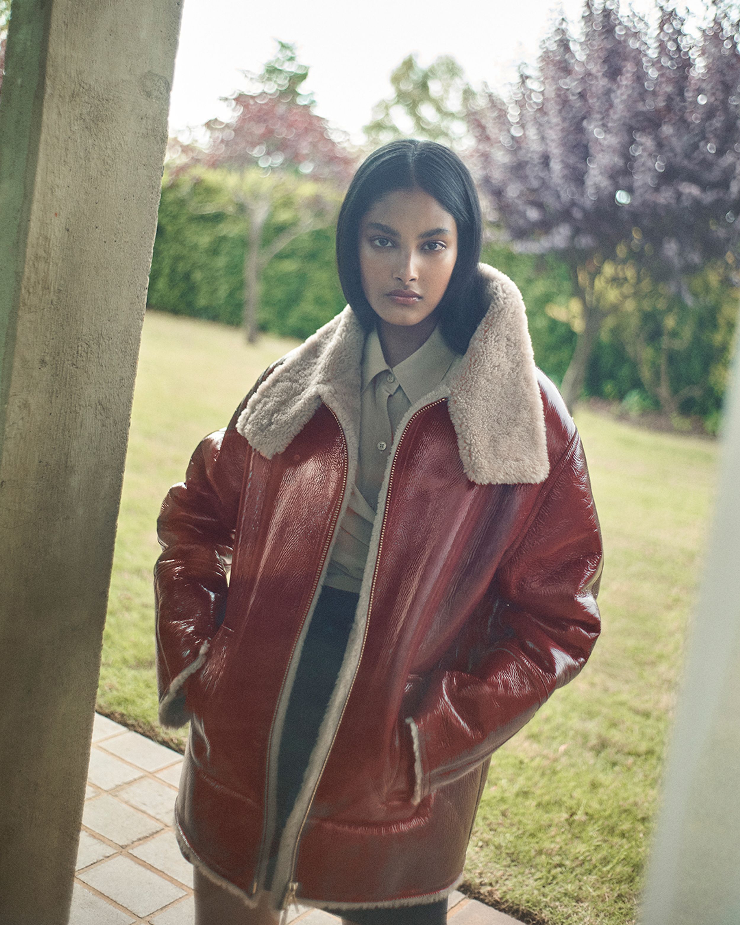 Brunette model standing in front of a garden wearing a red shearling coat