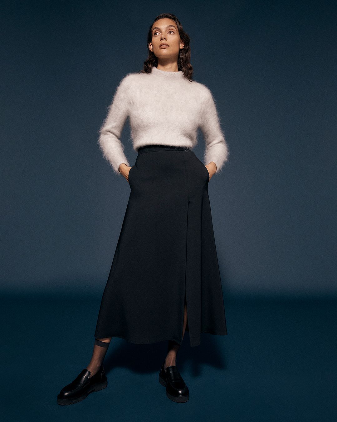 Brunette model standing with hands in pockets wearing a white mohair sweater and black skirt