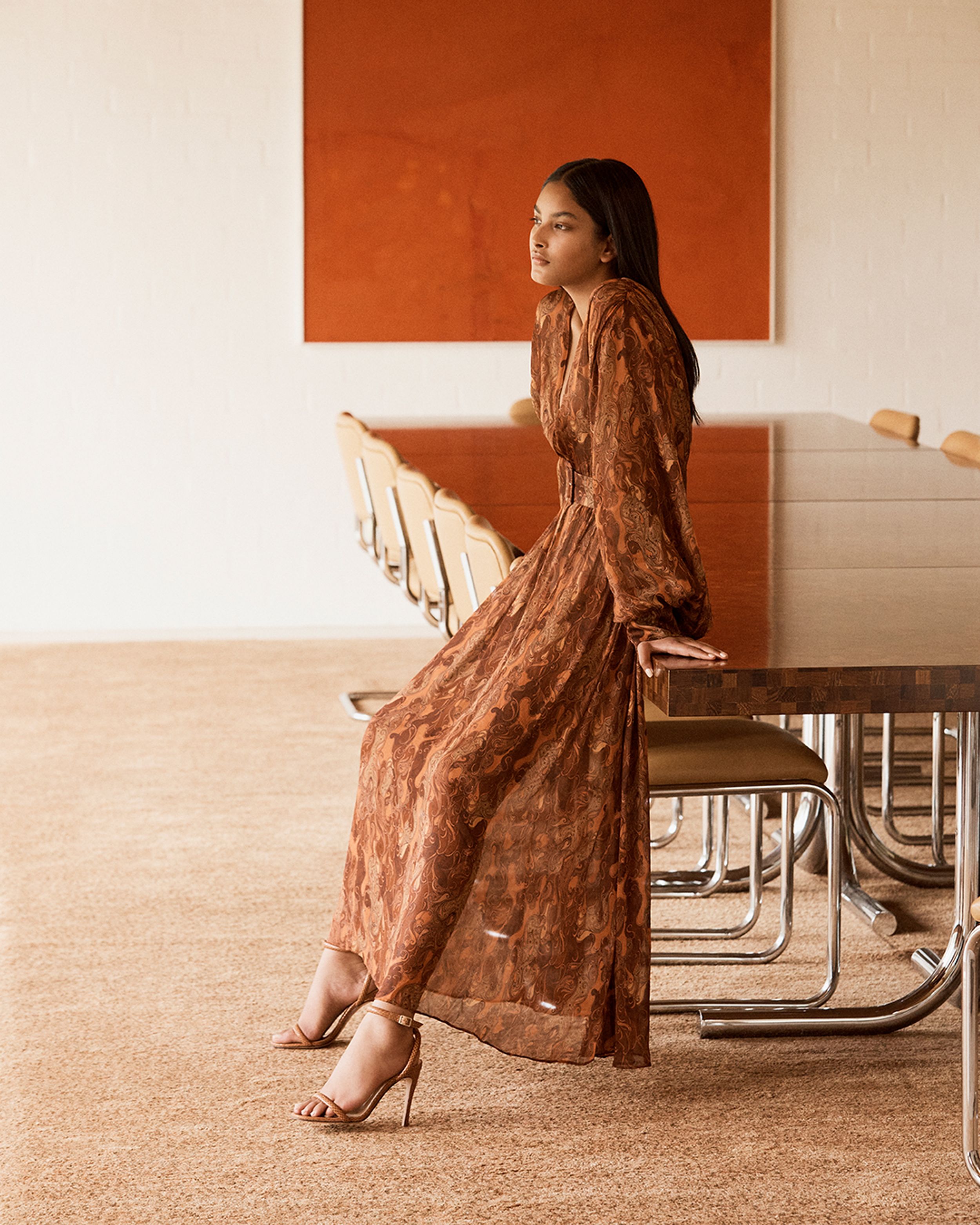 Brunette model leaning against a dining table wearing a brown print dress