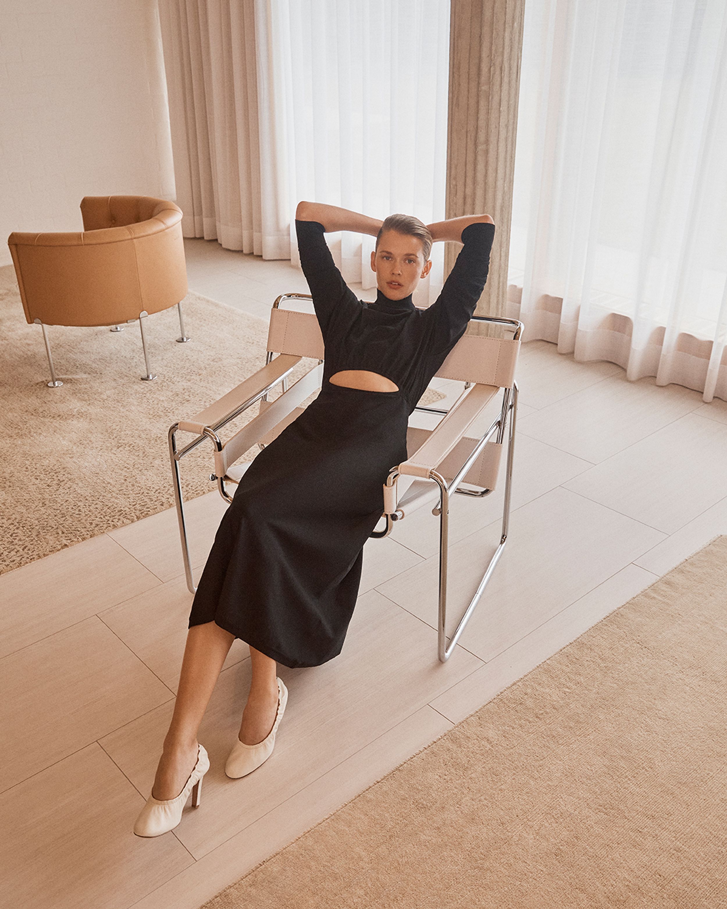 Model leaning back in arm chair with hands behind her head wearing a black long sleeve dress