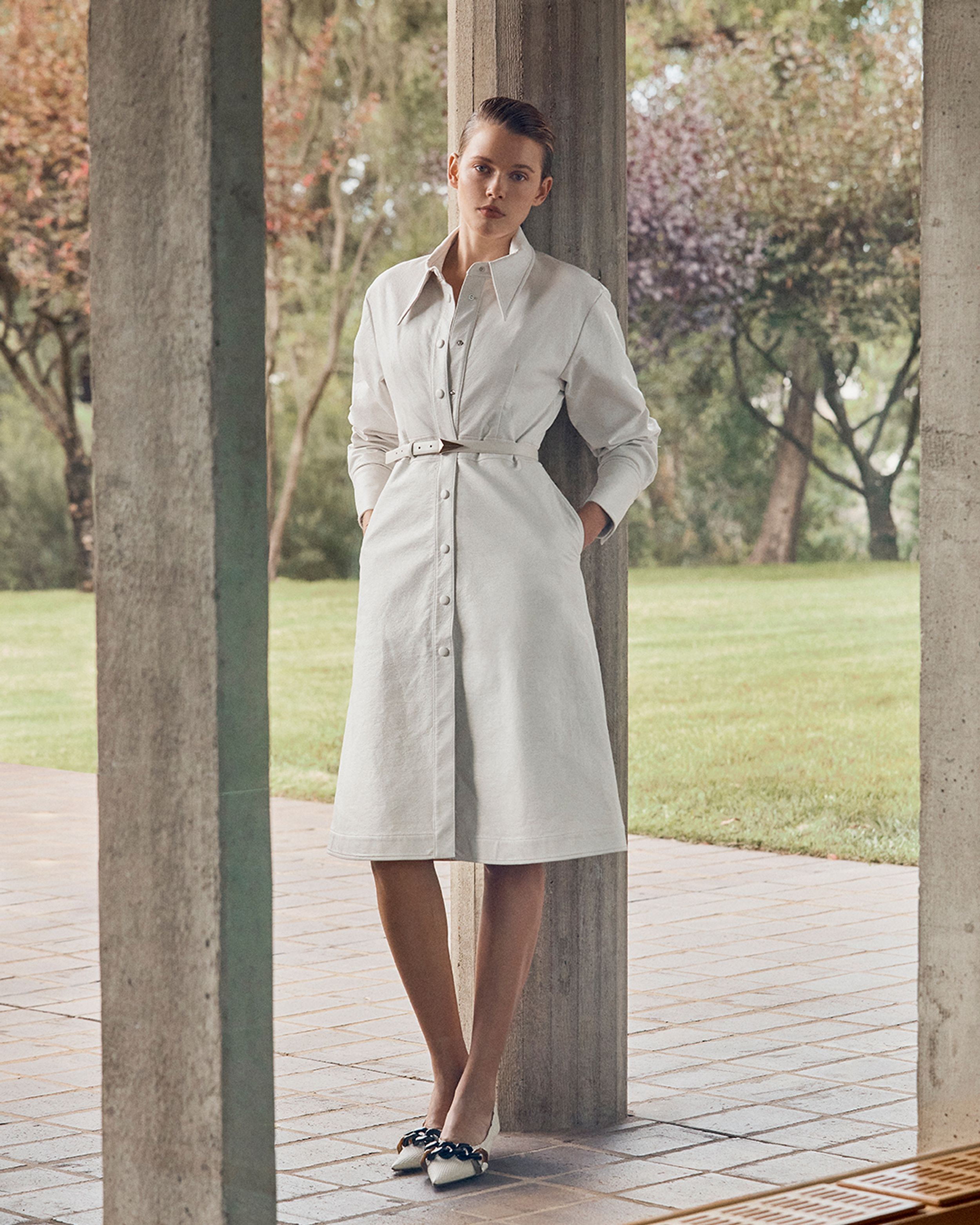 Model leaning against a pillar standing with a garden in the background wearing a white leather dress