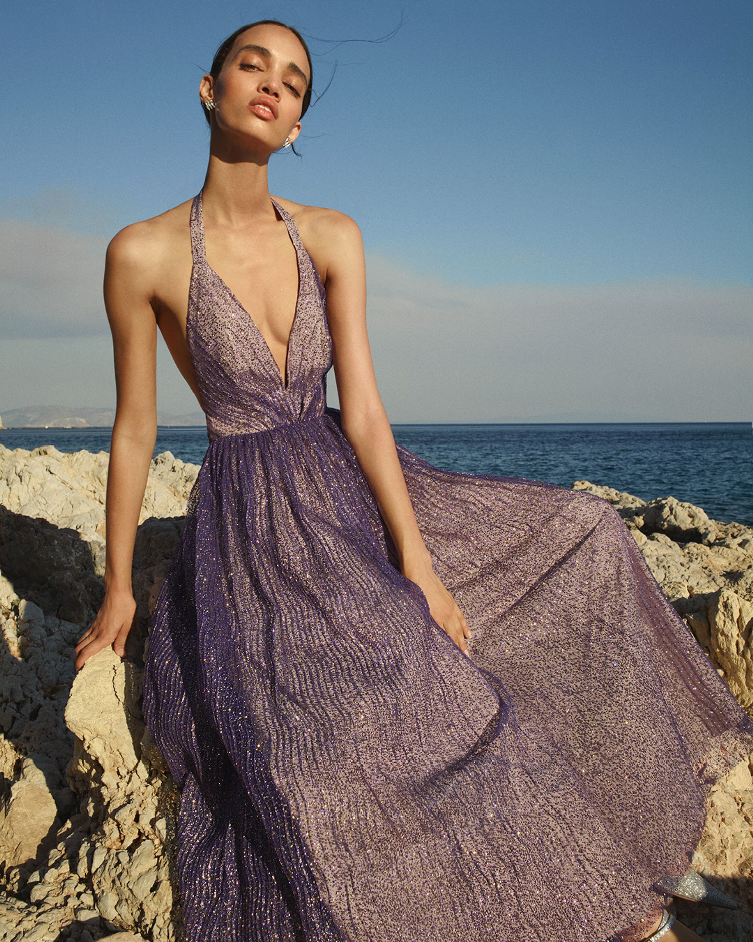 Model wearing a purple sparkly tulle gown leaning on rocks with the ocean in the background