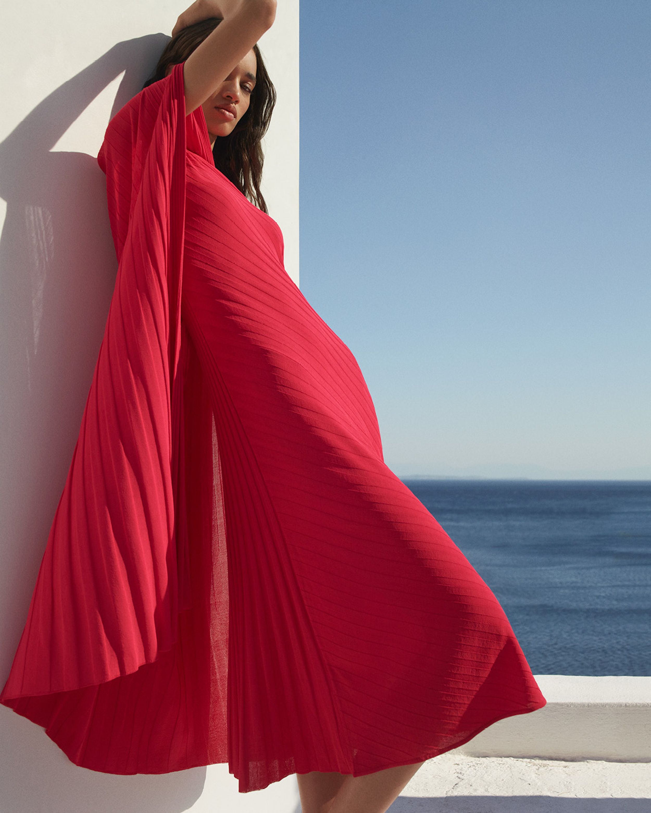 Model leaning against a wall with a hand on her hand wearing a bright pink dress with the ocean in the background