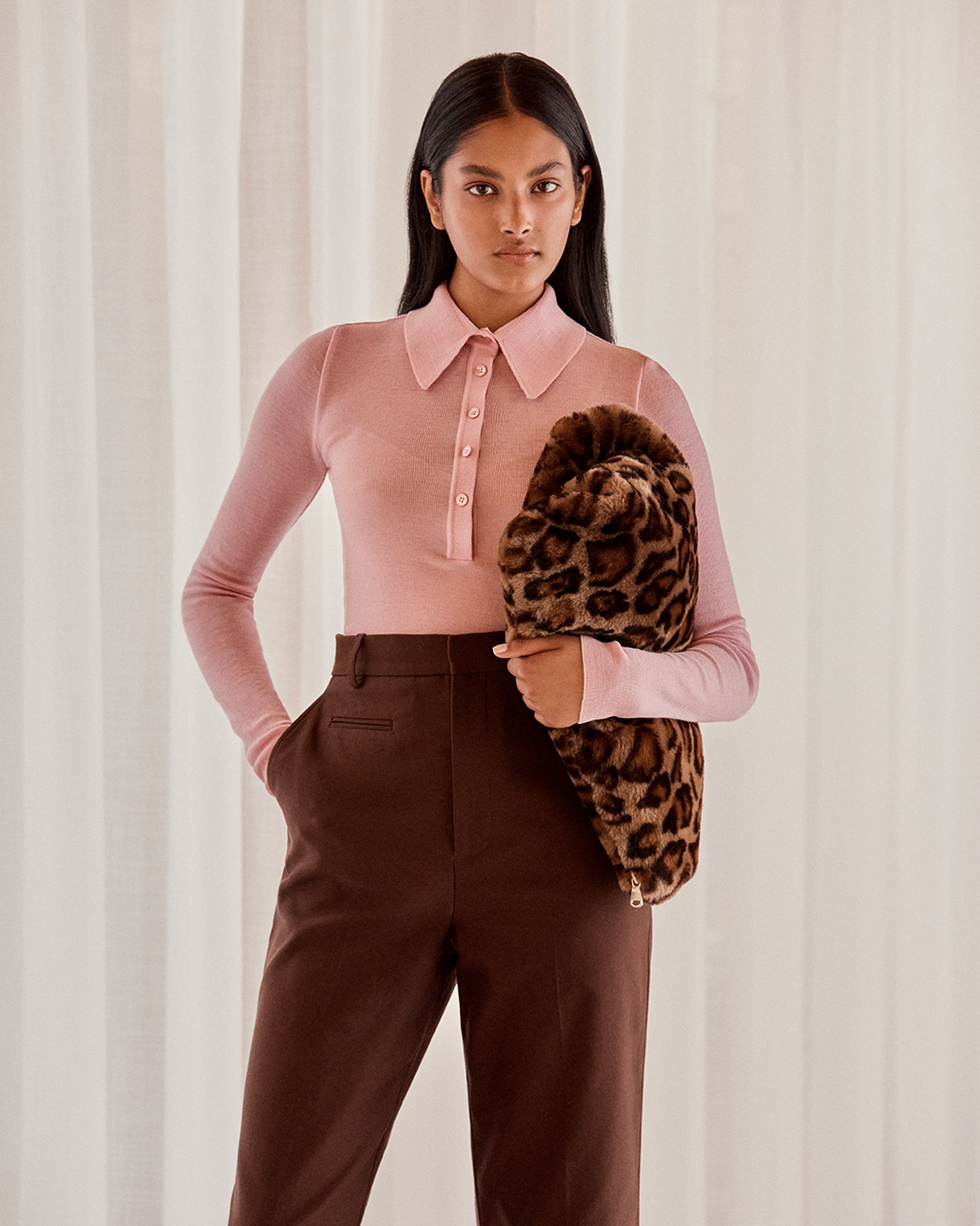 Brunette model standing by a window wearing a pink knit shirt holding a leopard-print clutch