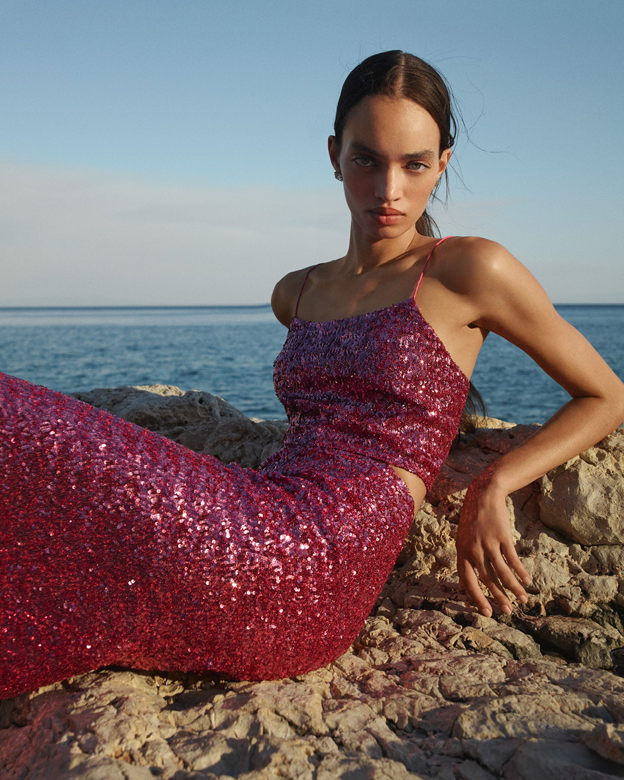 Model wearing a pink sequined singlet and skirt leaning on rocks with the ocean in the background