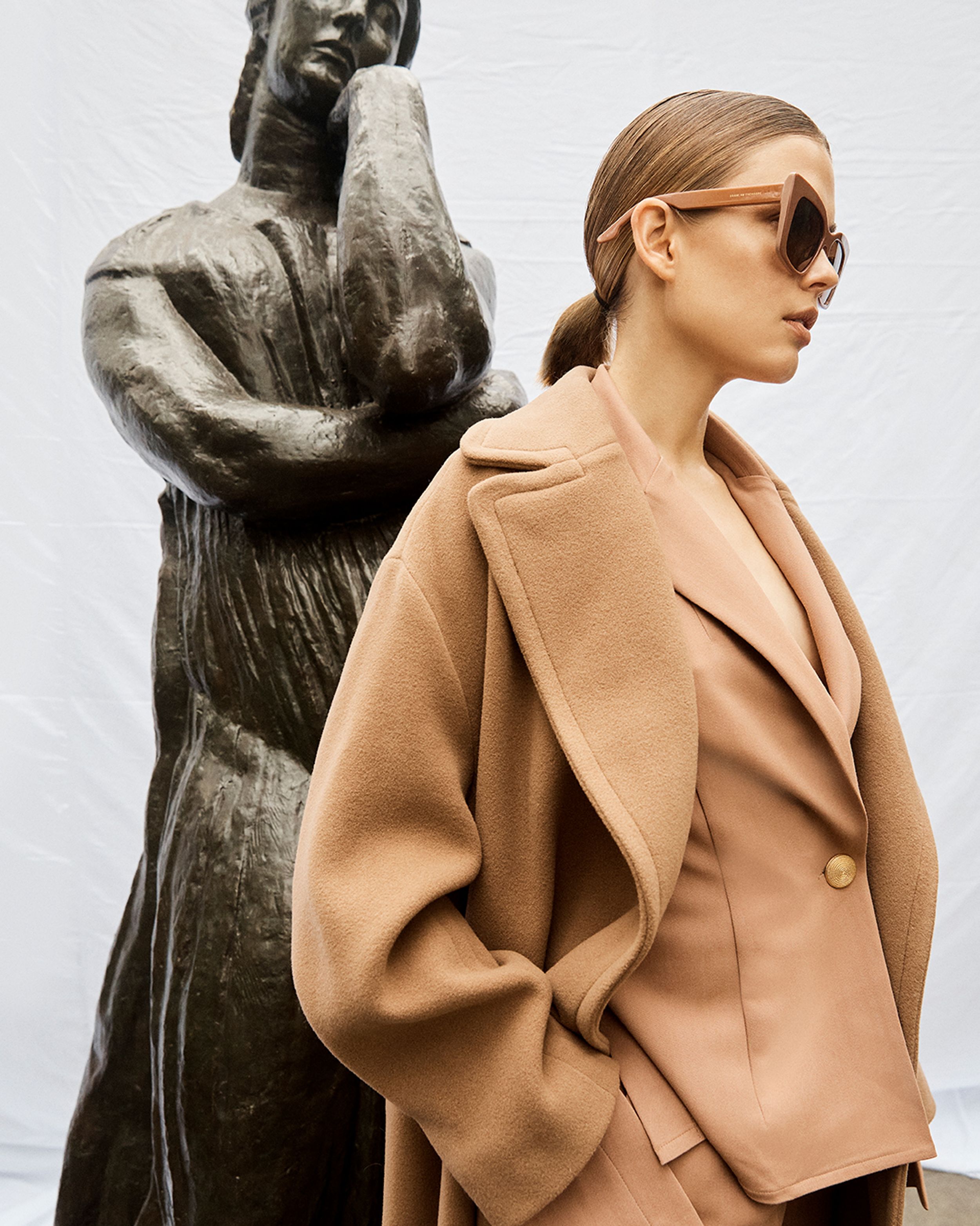 Model looking to the side wearing a camel-coloured coat next to a bronze sculpture