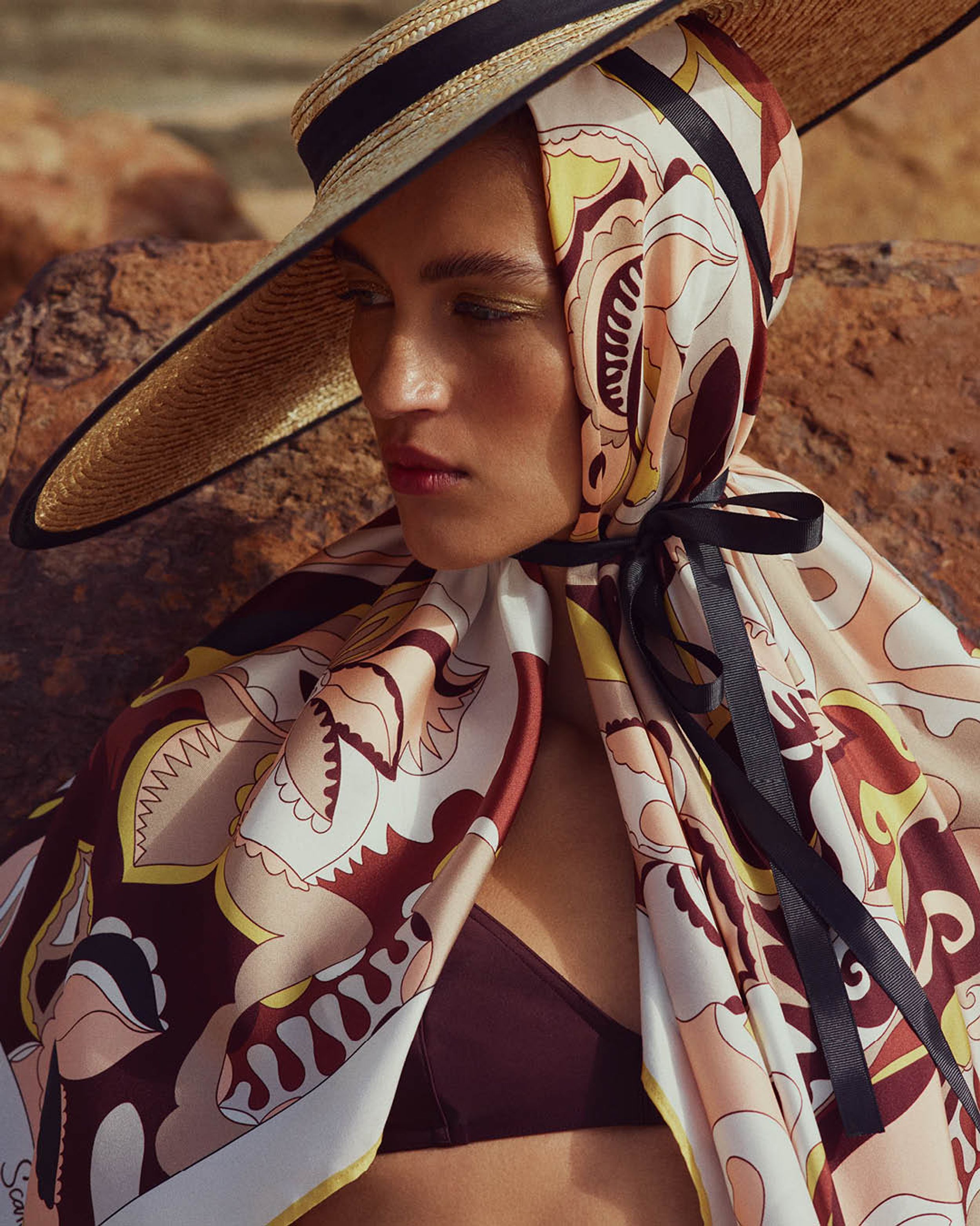 Model gazing downwards wearing straw hat with a brown, yellow and white scarf over her head and shoulders