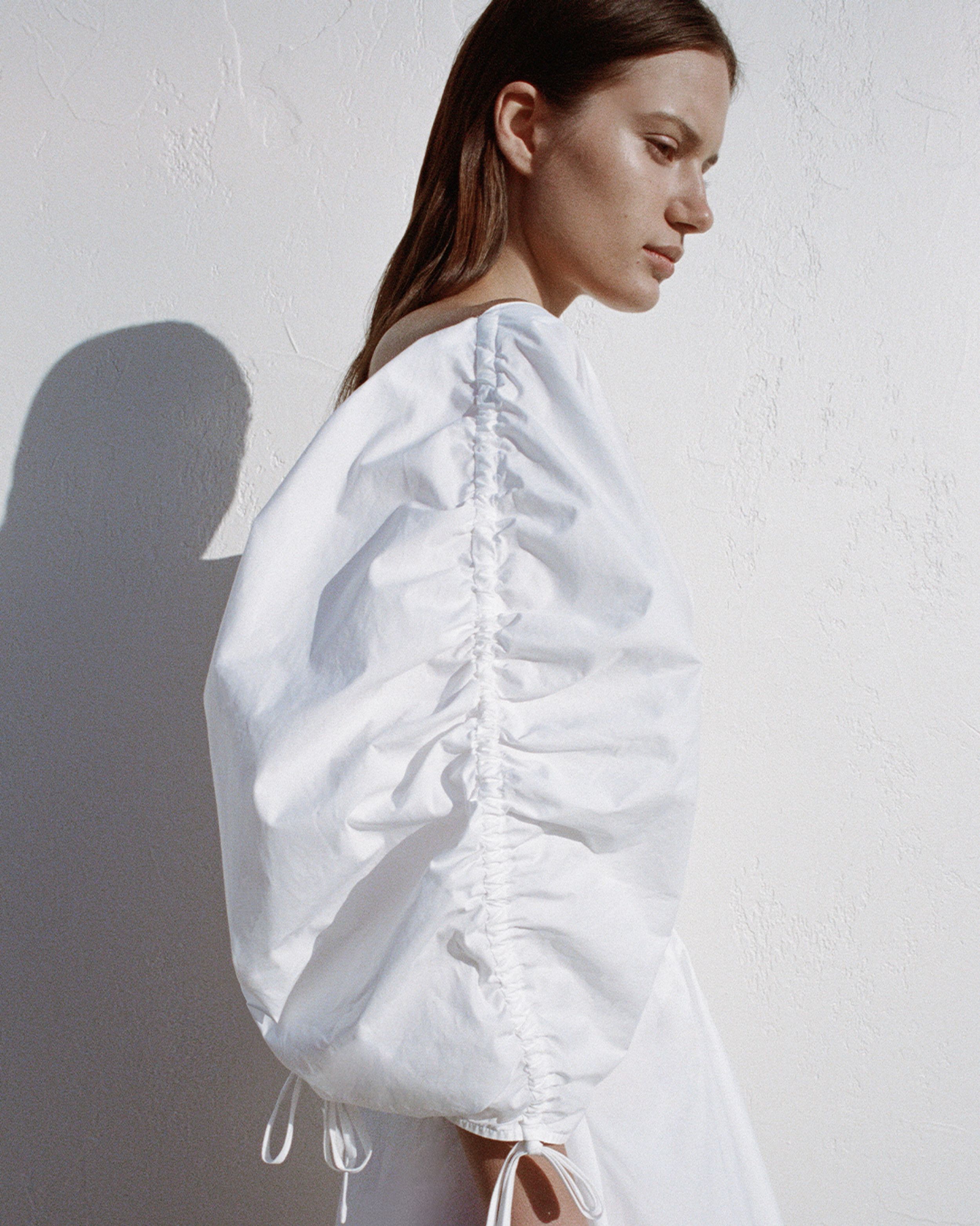 Profile photograph of brunette girl showing white dress sleeve, with shadow on white wall. 