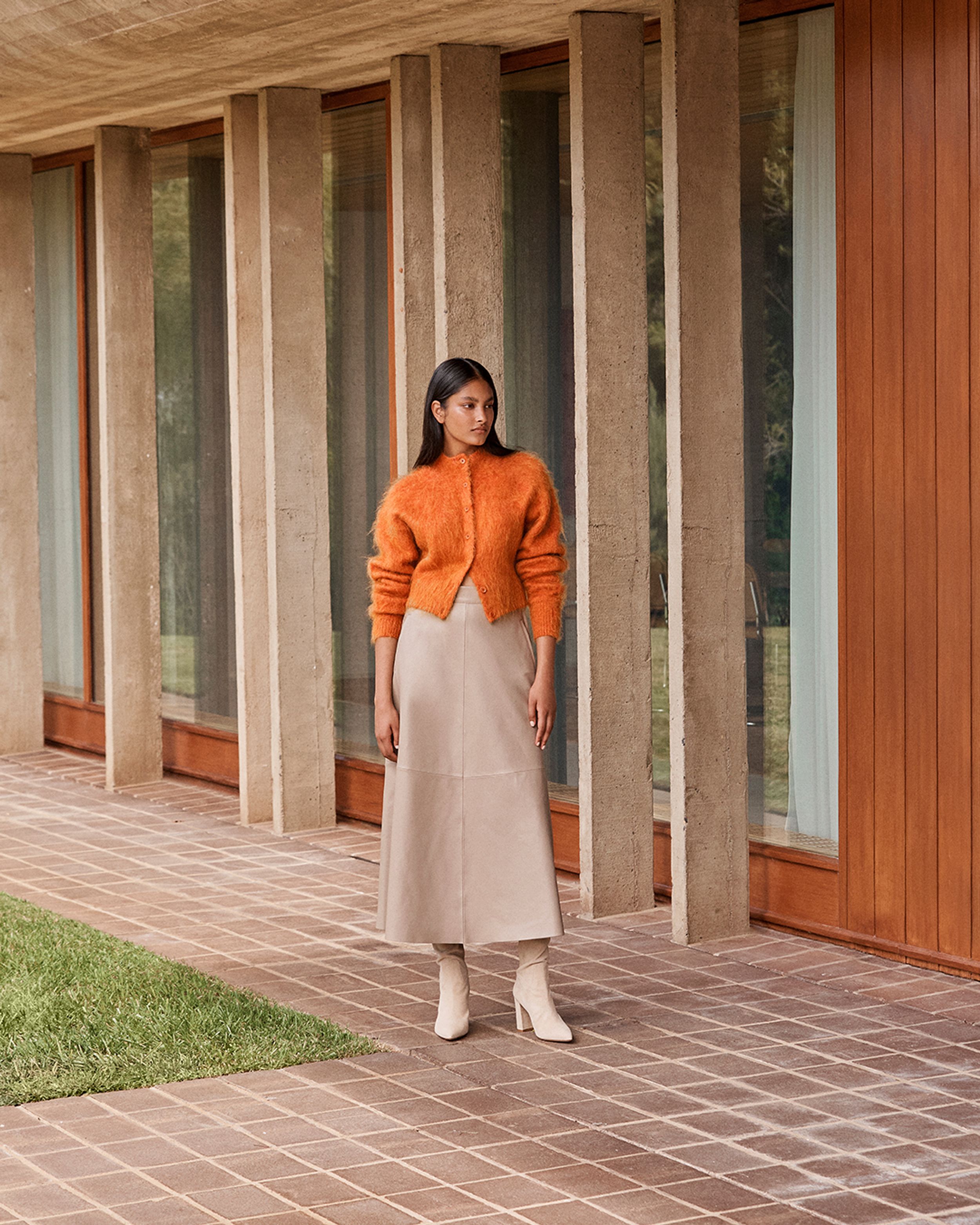 Brunette model standing outside wearing orange knit cardigan and taupe leather skirt