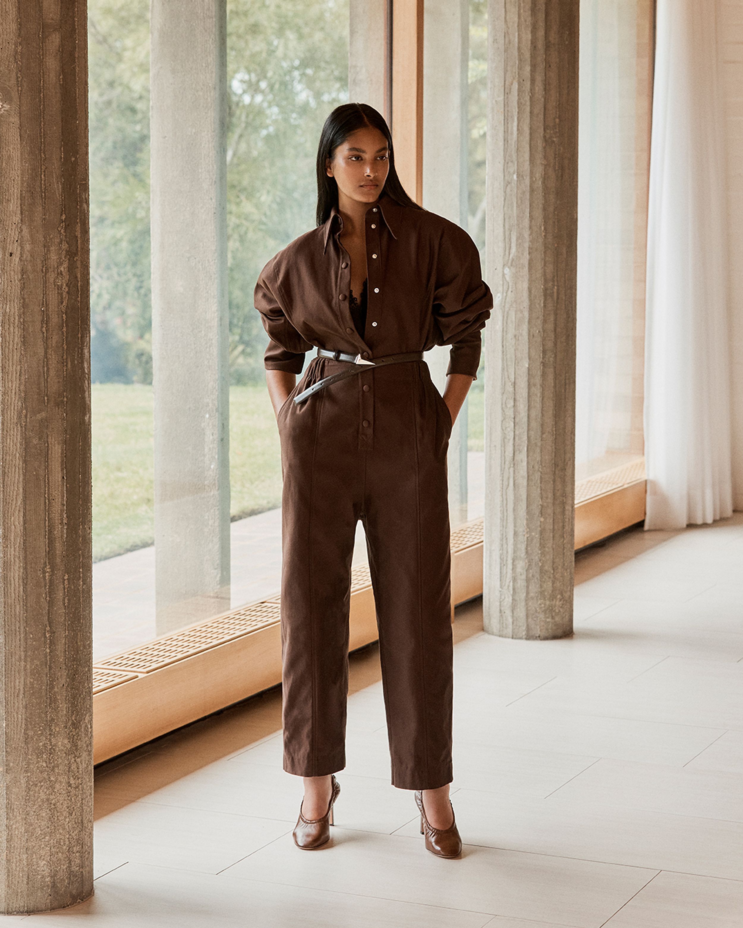 Brunette model standing by a window wearing a brown jumpsuit with belt