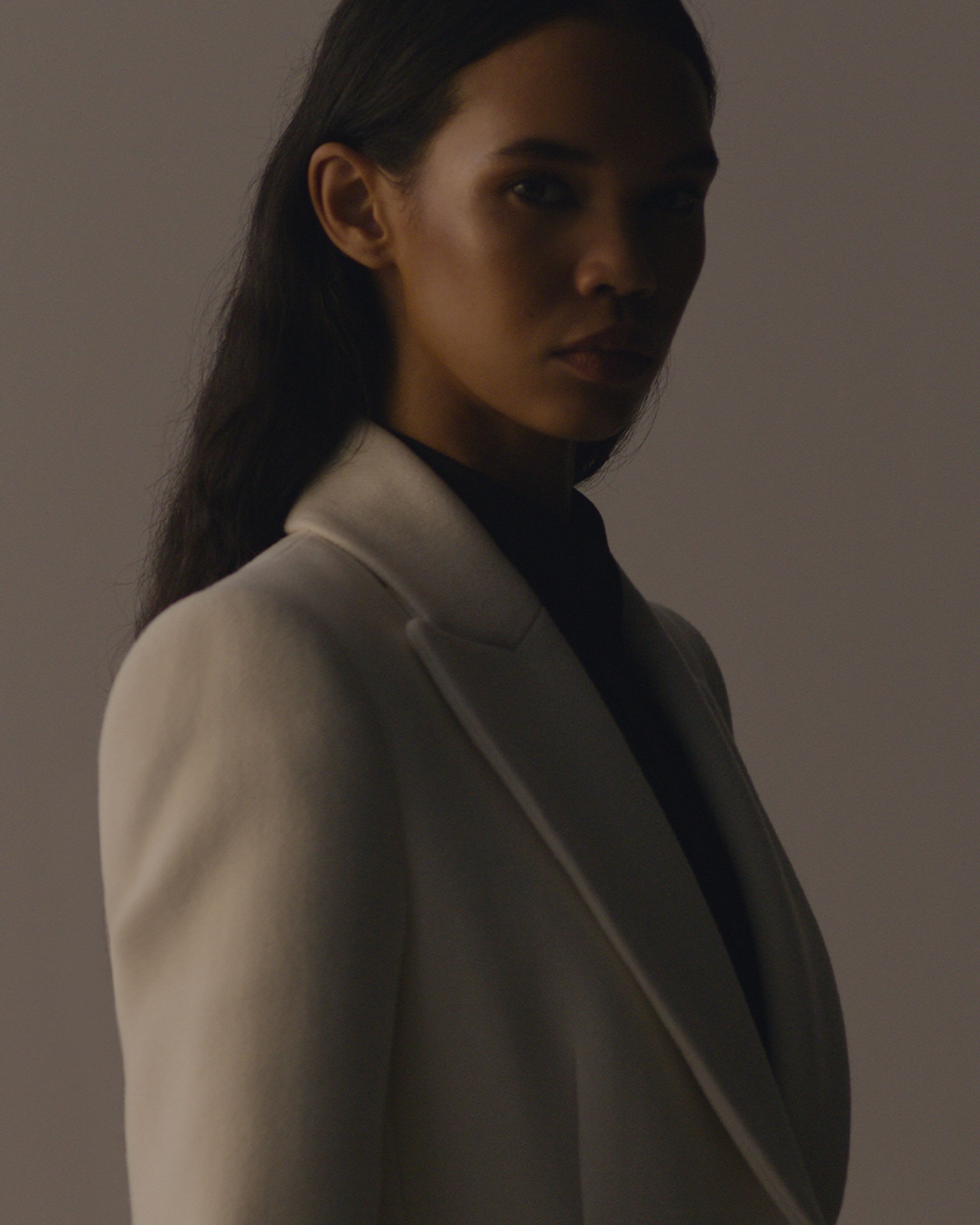 Women standing side on looking at camera in darkness, hair tucked behind ear white coat with lapel. 