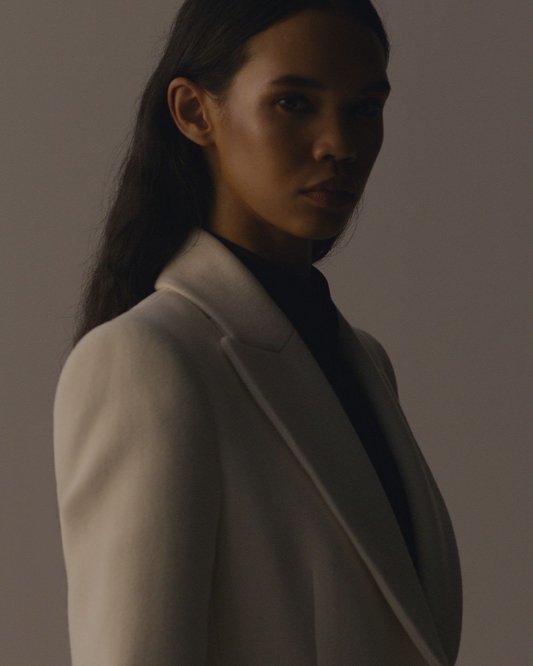Women standing side on looking at camera in darkness, hair tucked behind ear white coat with lapel.