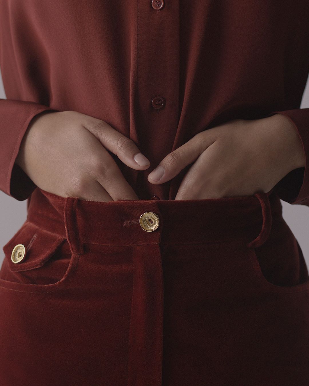 Woman’s hands tucking into red velvet trousers, gold button on front of pants and pocket.