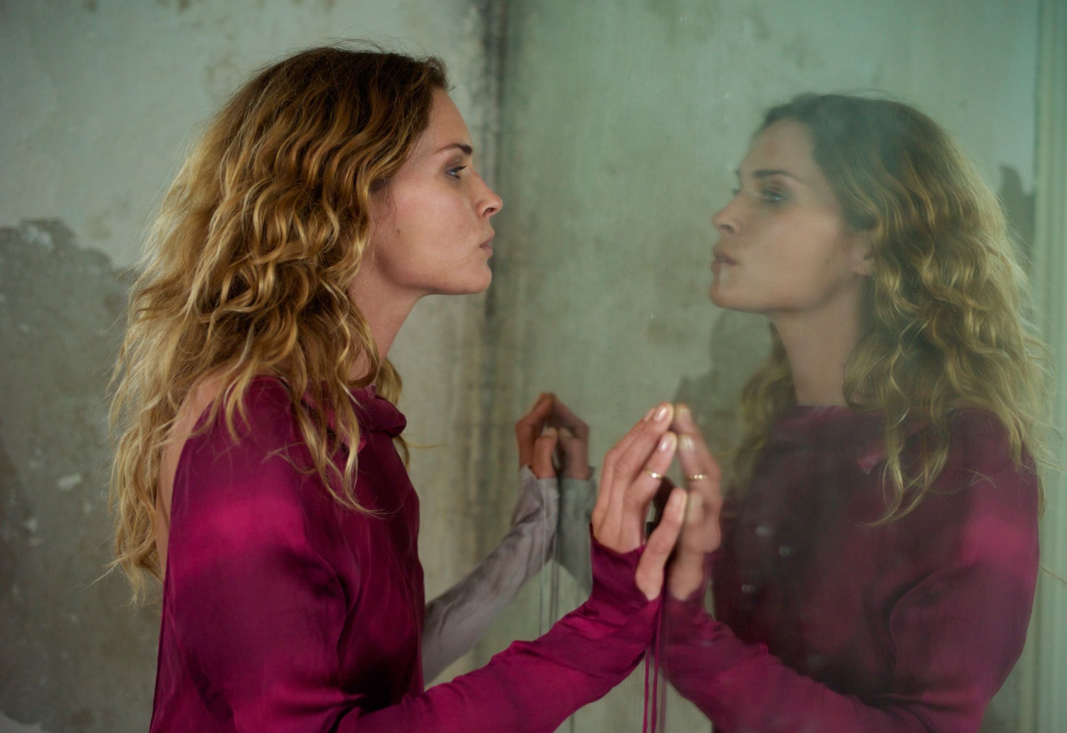 Erin Wasson looking into a mirror with her hands placed on it, wearing a raspberry pink long-sleeved top