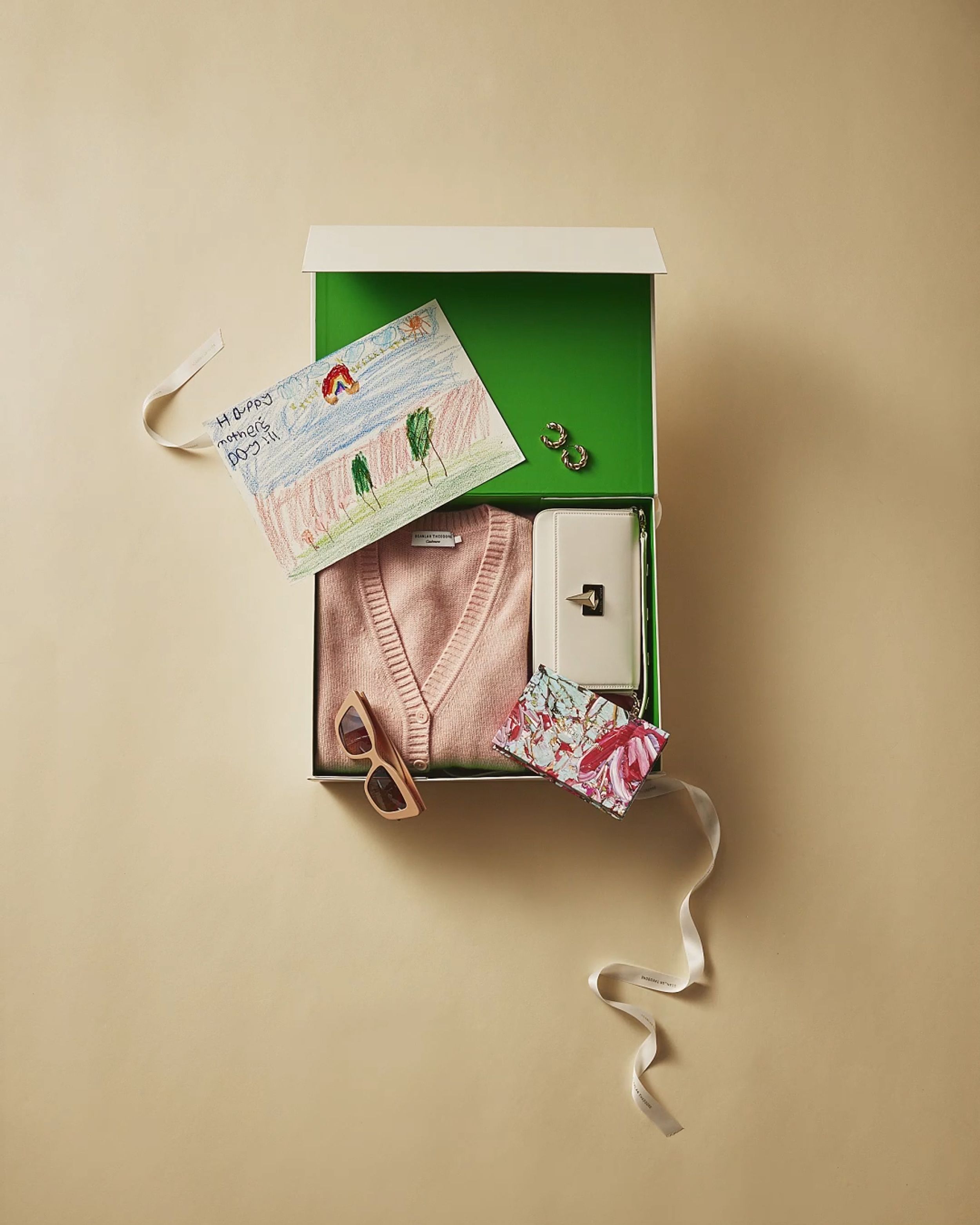 Flatlay of an open green gift box with pink cardigan, sunglasses, white bag, gift card and child's drawing