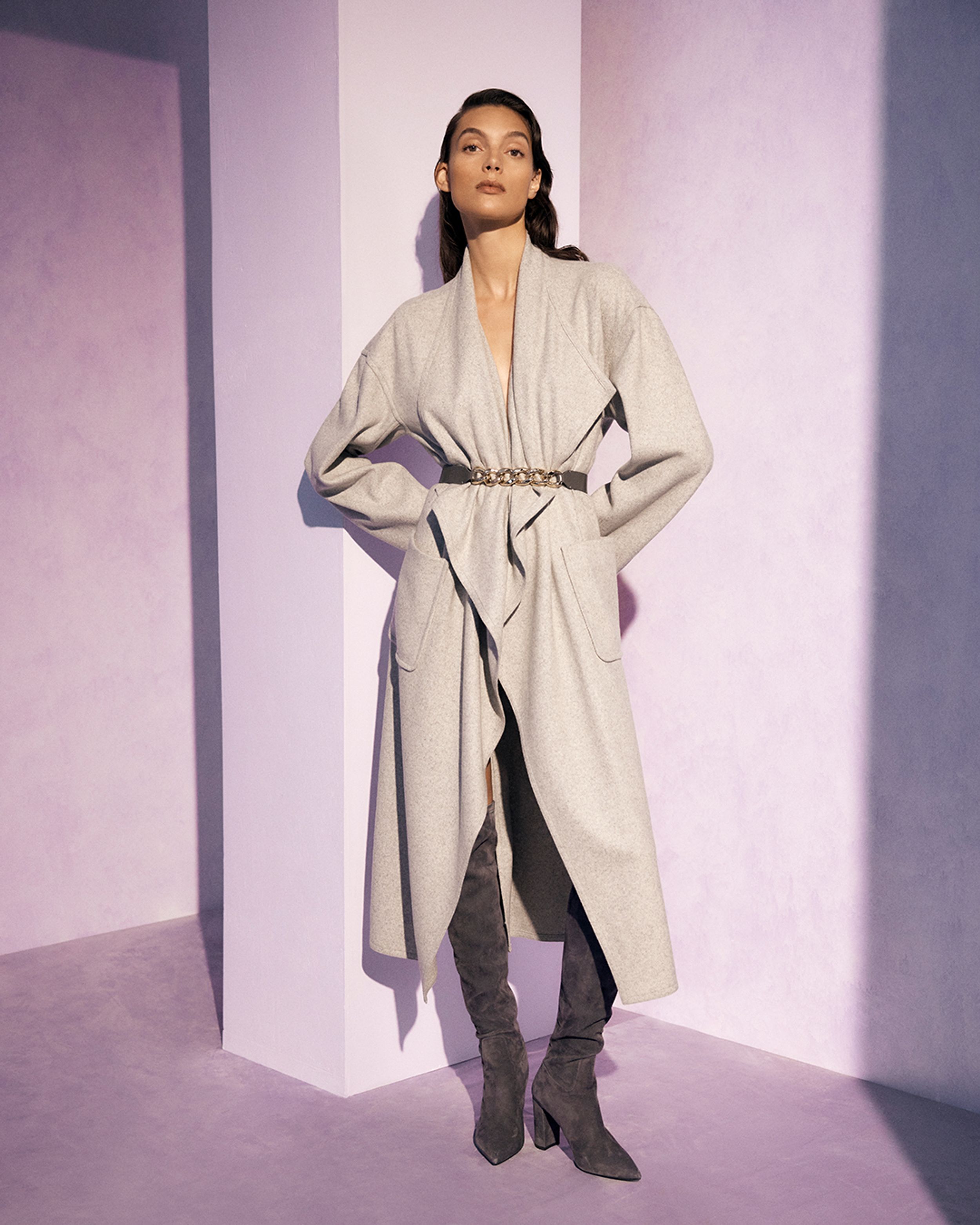 Brunette model standing with hands on hips in a neutral-toned coat with grey over-the-knee boots