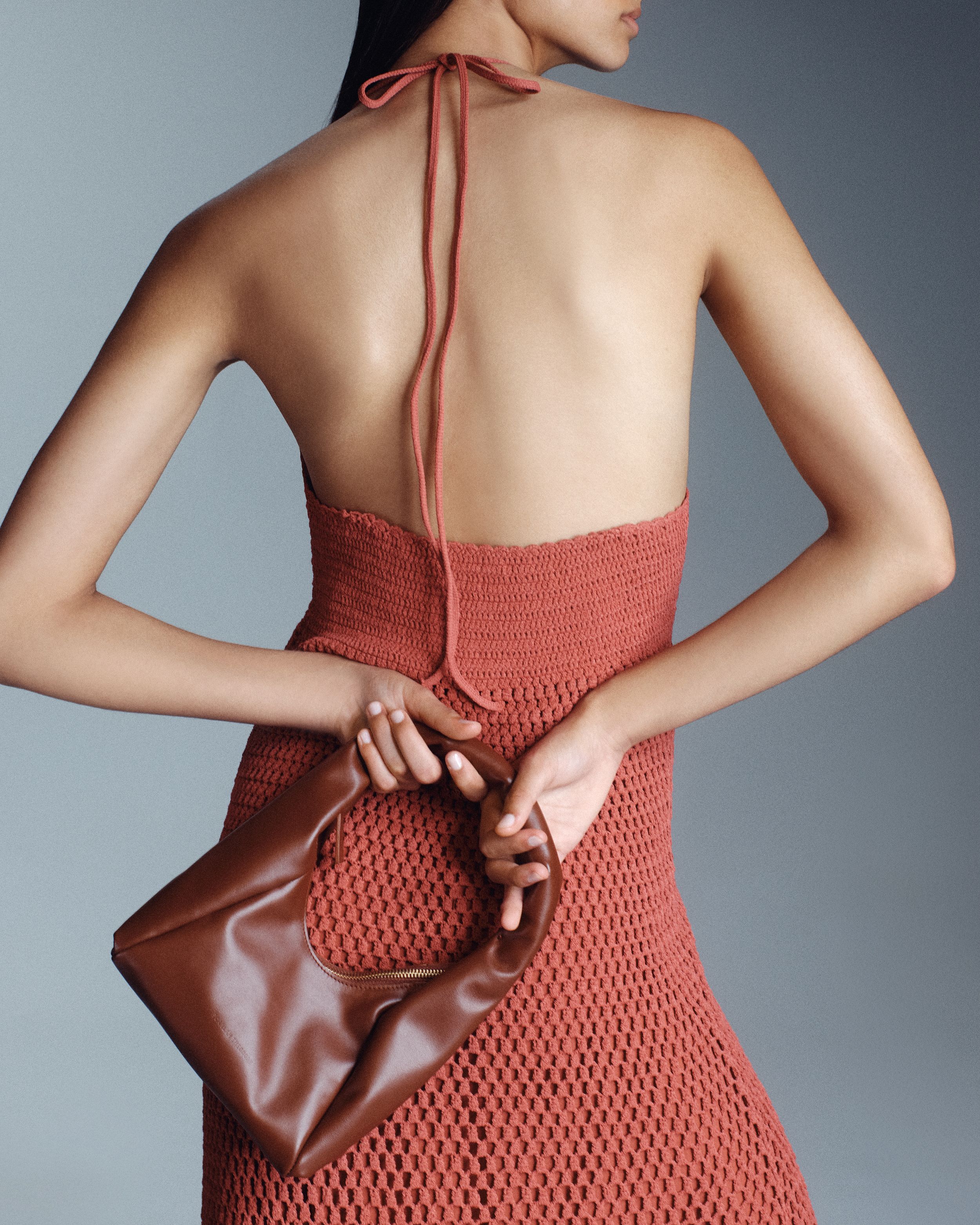 Model facing the wall holding a small brown leather bag behind her black wearing a orange crochet halterneck dress