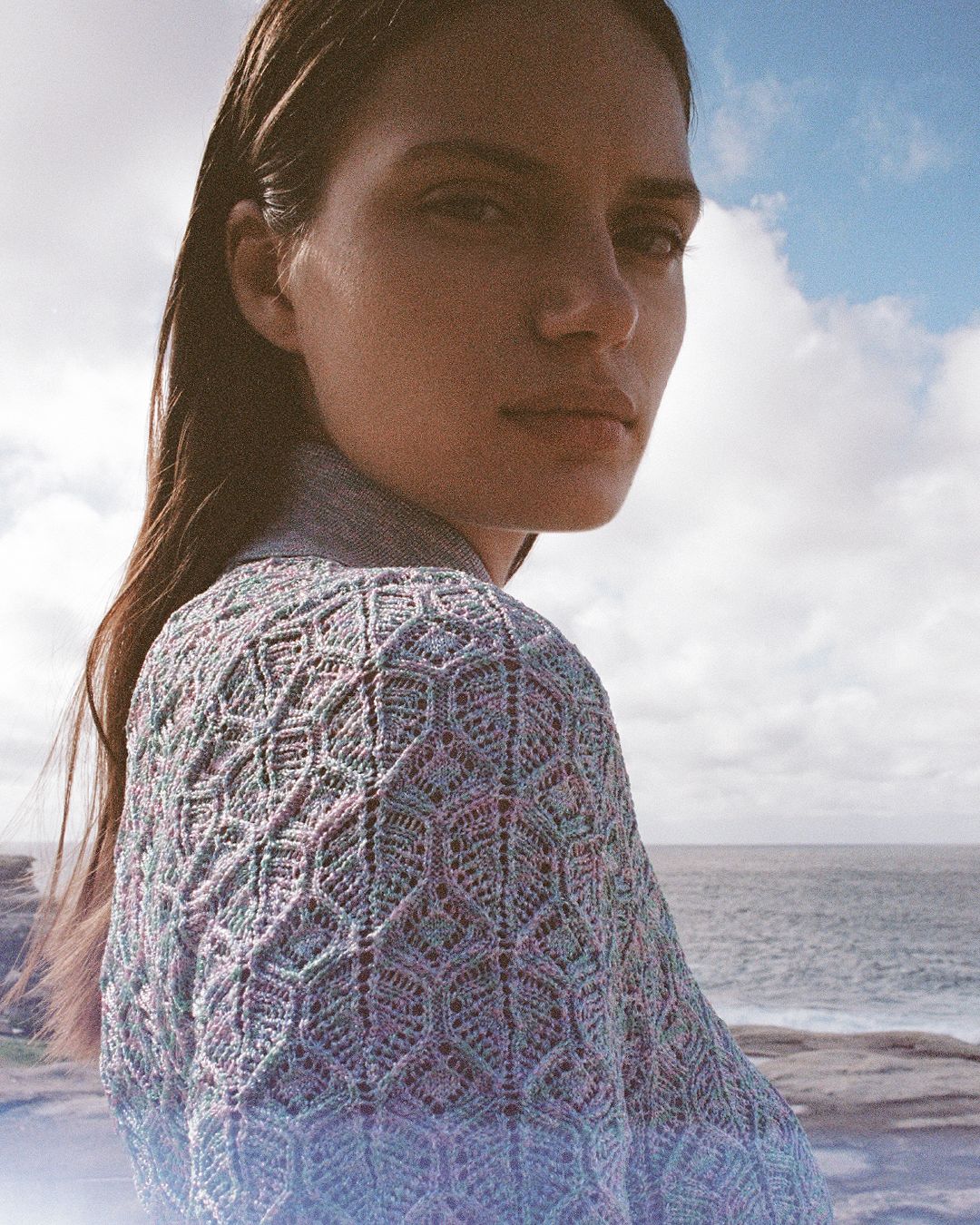 Girl standing side on looking at the camera in blue knit dress with sea and clouds in background.