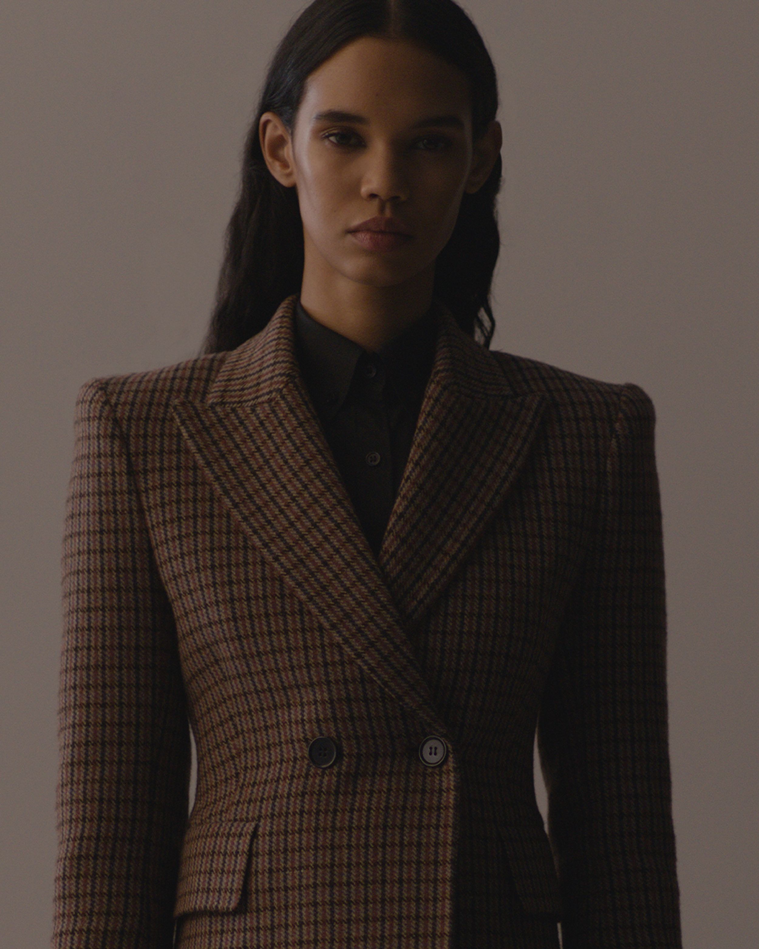 Woman wearing red and brown plaid blazer with shoulder pad dark lighting shadowing right side face. 