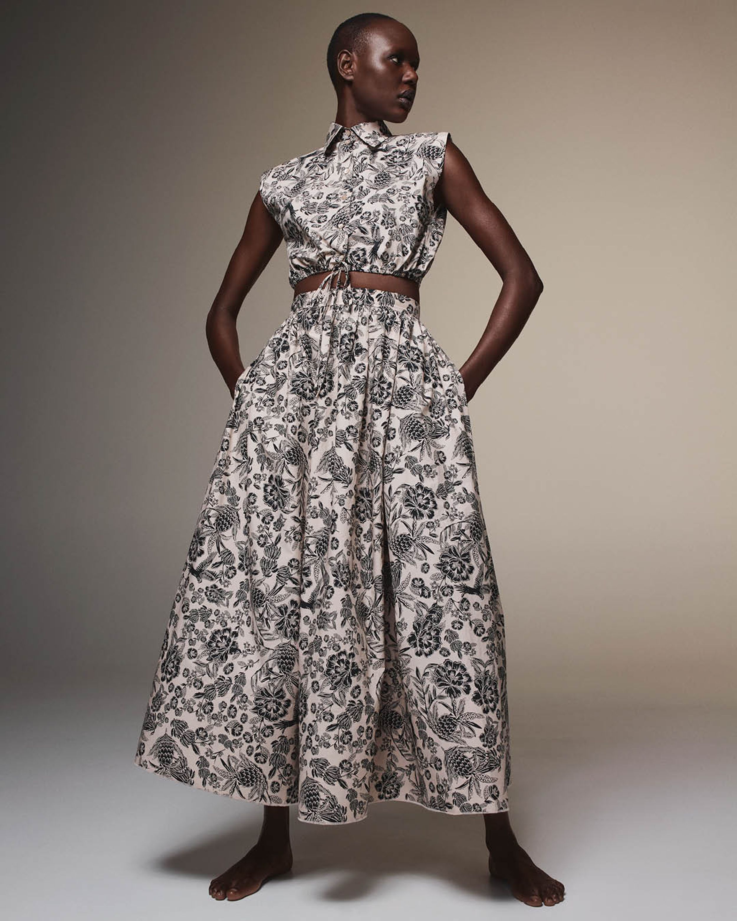 Ajak Deng standing with hands in pockets and head turned wearing a matching grey floral cropped shirt and long skirt