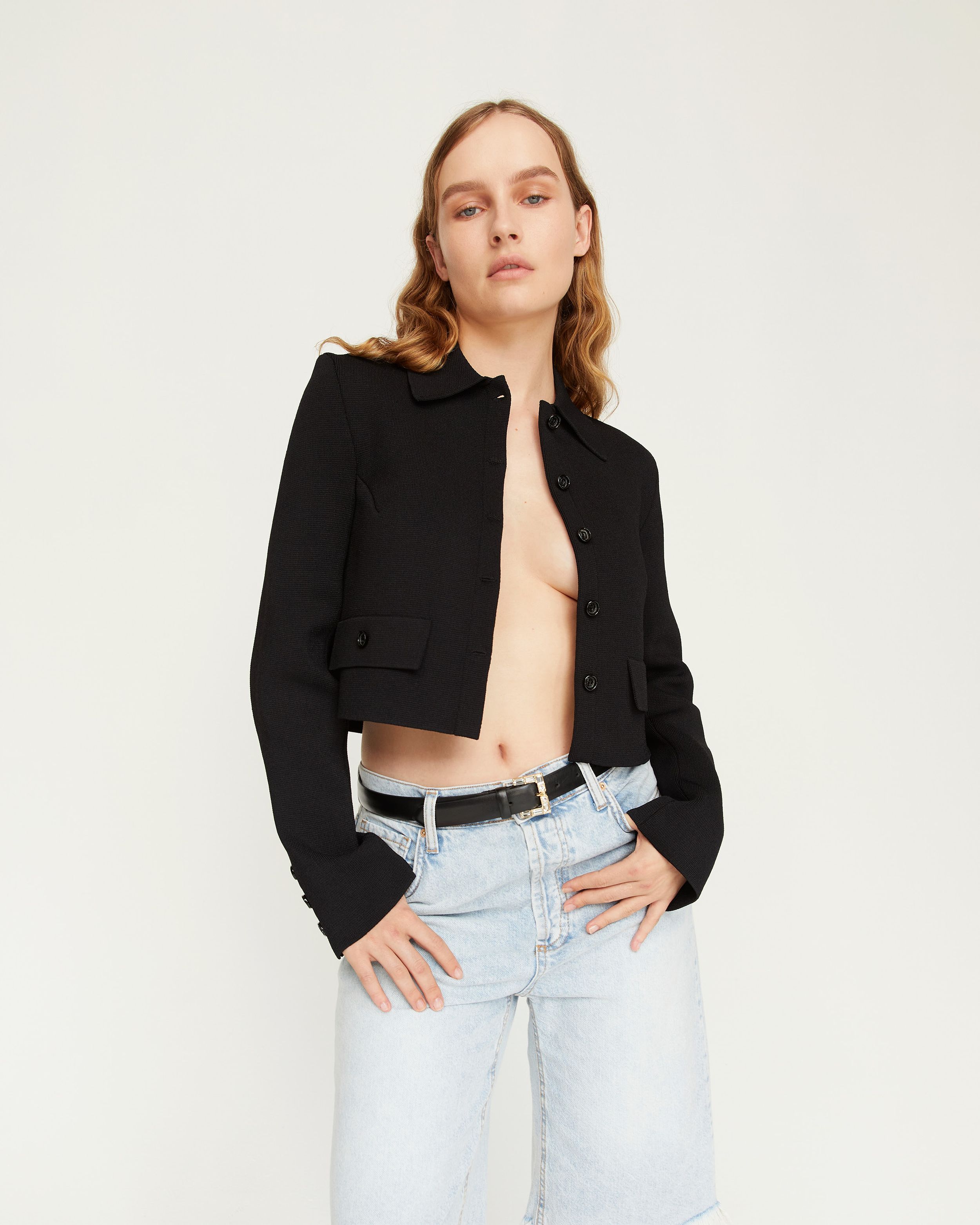 Olivia DeJonge with hands resting on pockets, wearing a black cropped jacket with bleach denim shorst