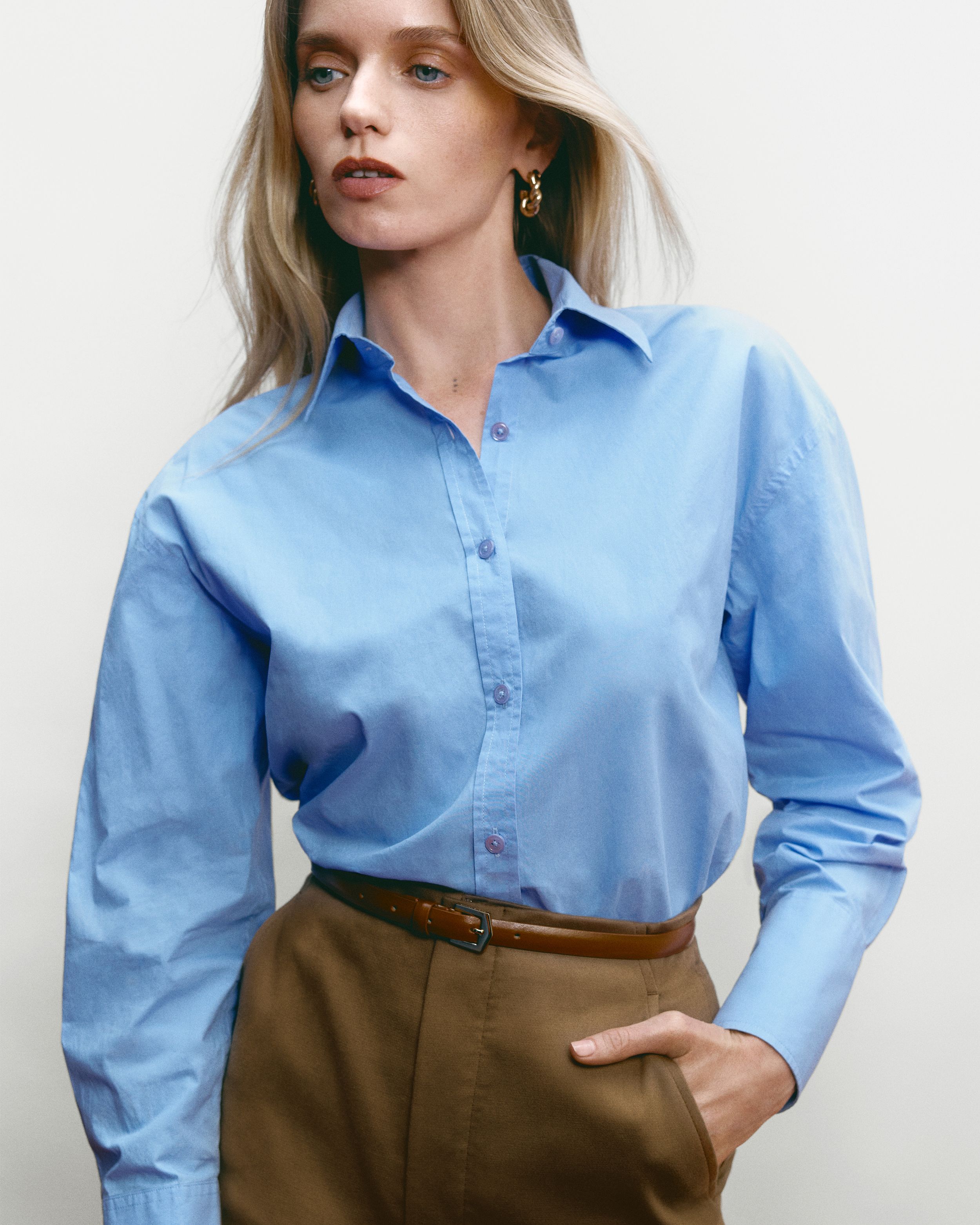 Abbey Lee standing with one hand in pocket wearing a blue cotton shirt and tan trousers