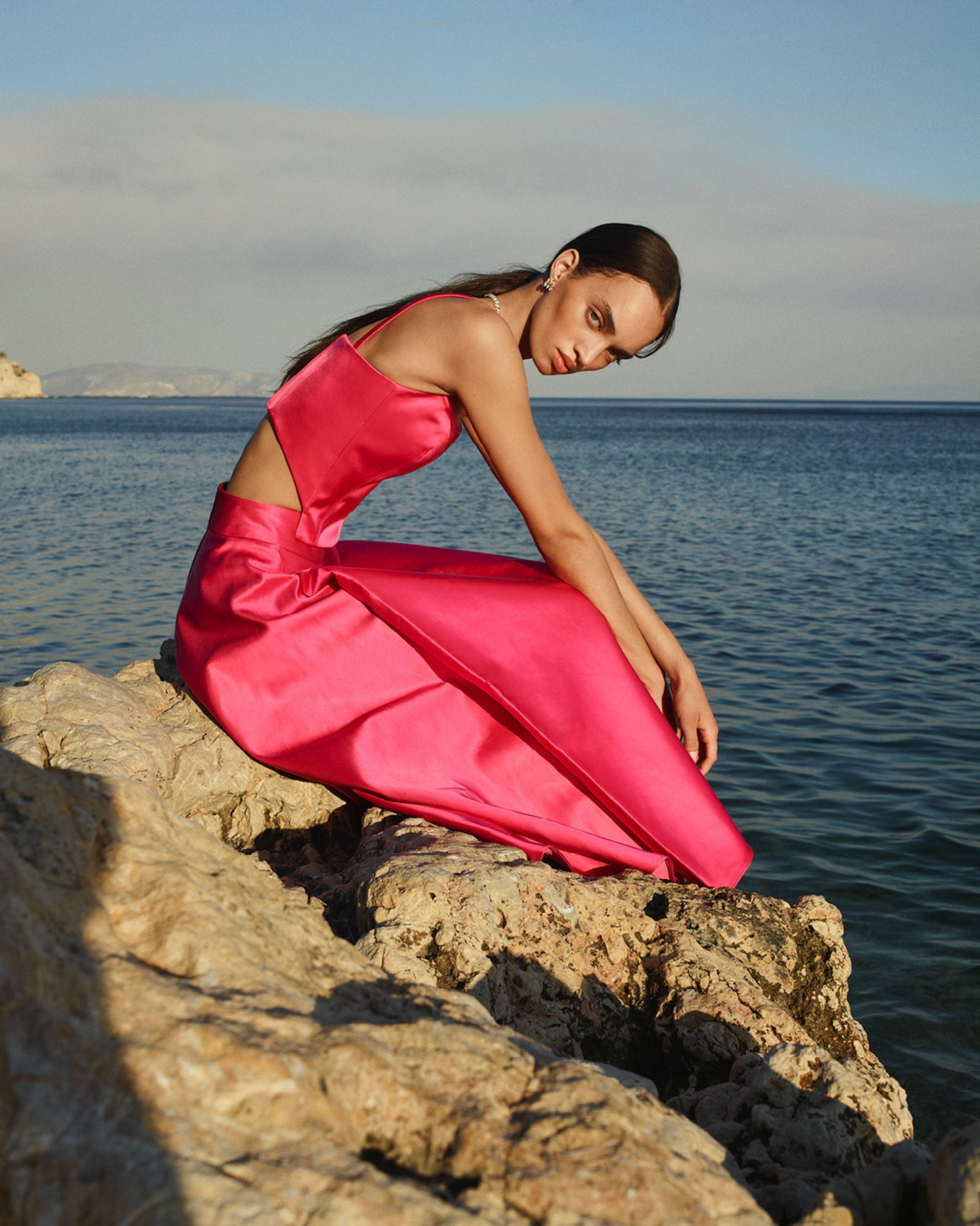 Model sitting on rocks in front of the ocean wearing a matching fuschia satin bustier and high-waisted skirt