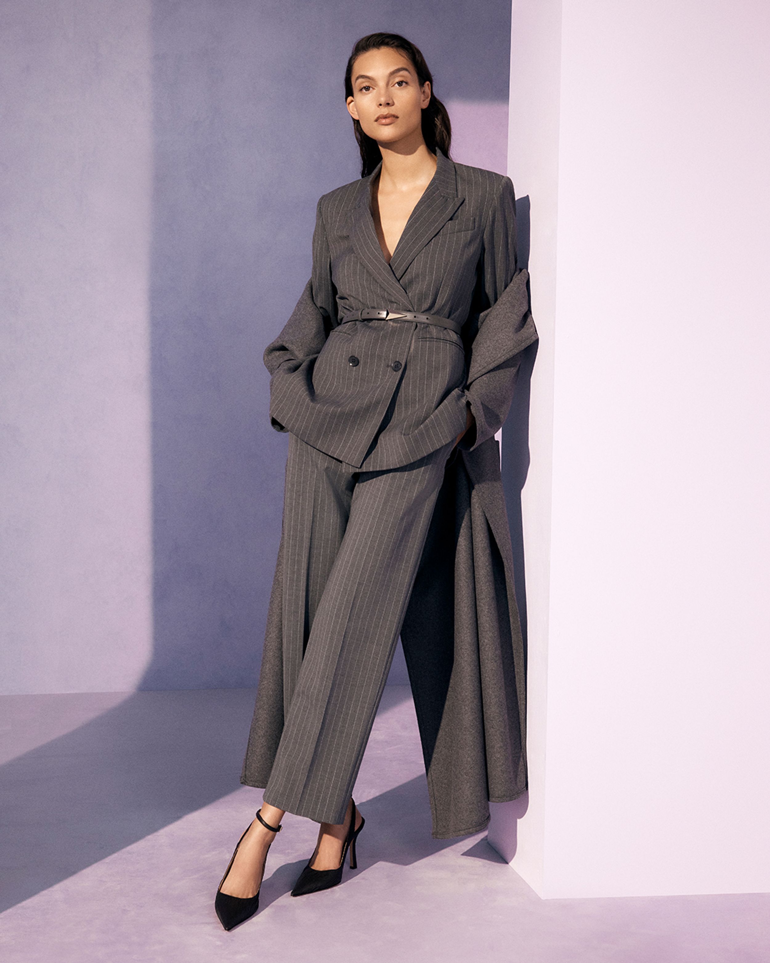 Brunette model learning against the wall wearing a charcoal pantsuit