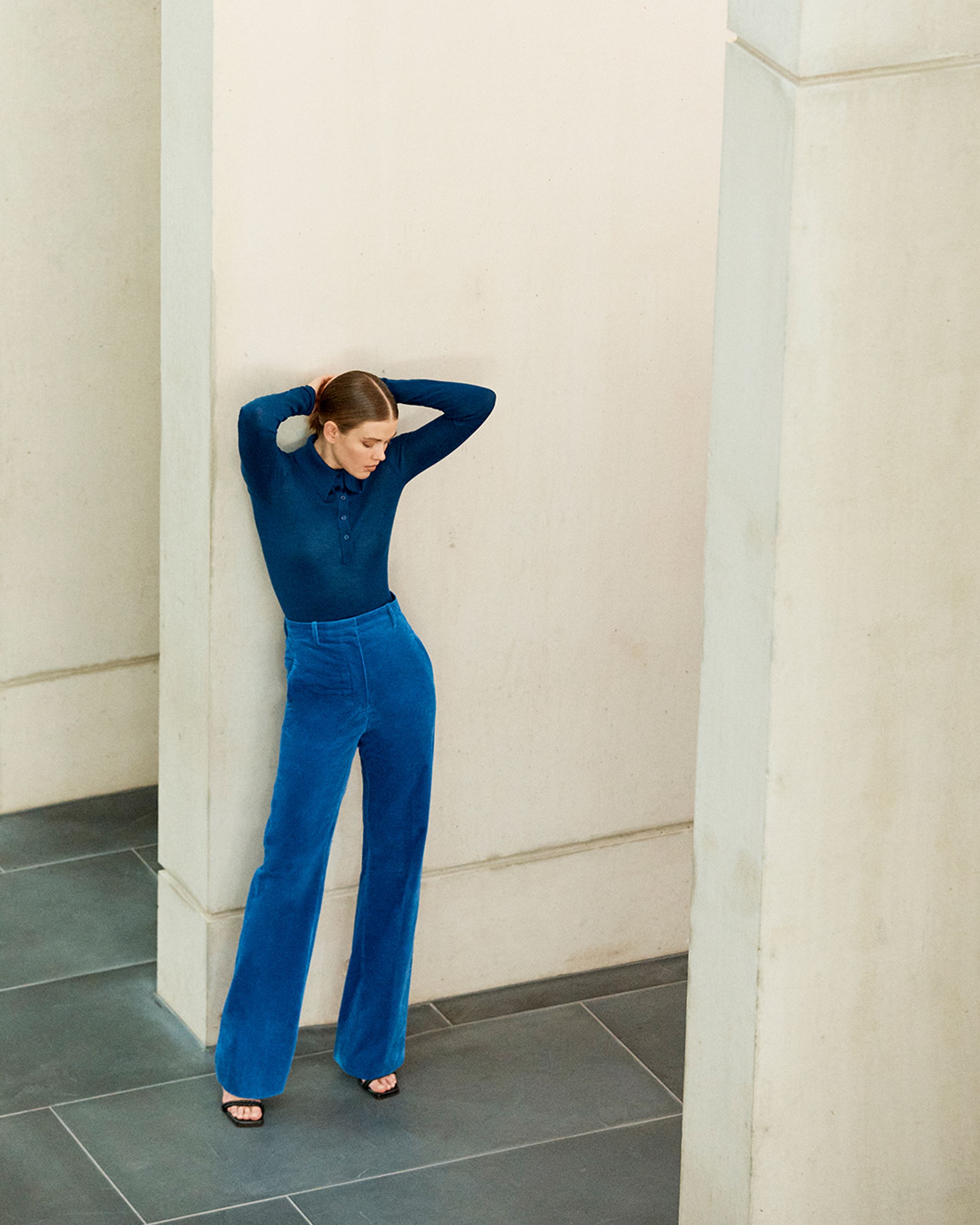 Brunette model leaning against a wall a blue babywool top and blue velvet trousers