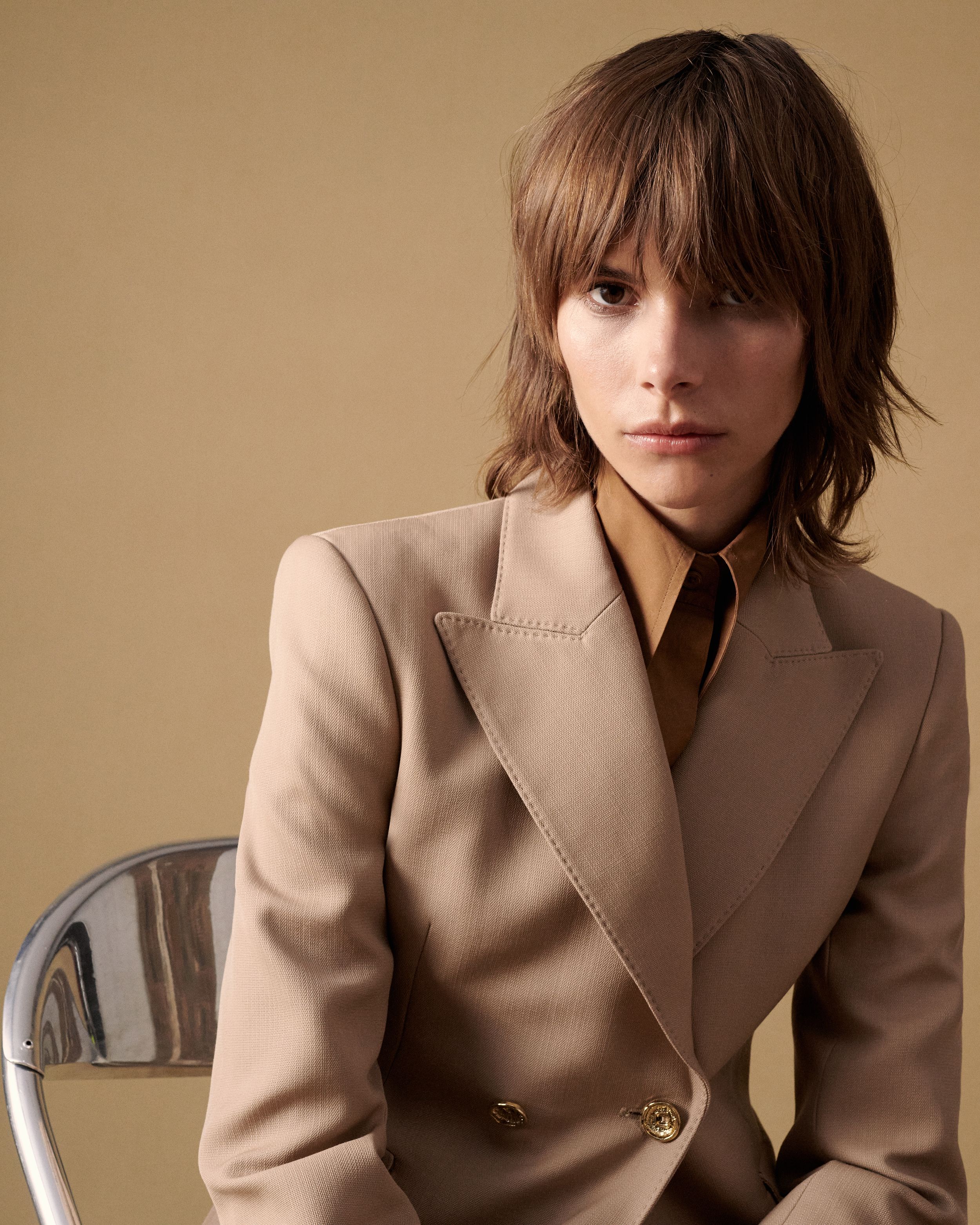Model sitting in a chair wearing a neutral-toned jacket and collared shirt